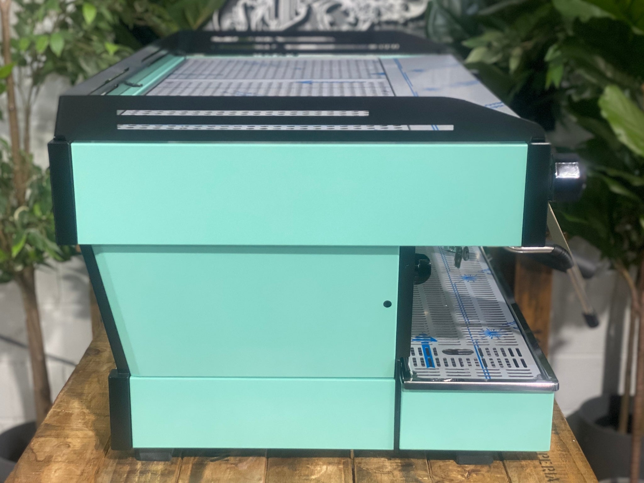 La Marzocco Linea PB 3 Group Coffee Machine Aqua - New espresso machine photographed from the side, showcasing the aqua side panel, body shape, and steam wand positioning on a wooden bench with greenery.