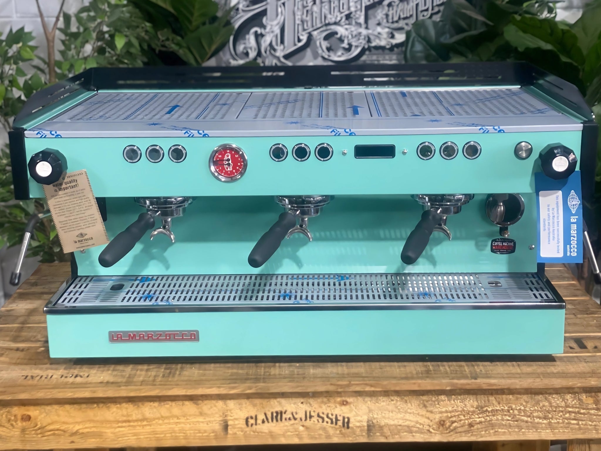 La Marzocco Linea PB 3 Group Coffee Machine Aqua - New espresso machine photographed front-on with the top panel visible, showing the group heads, cup tray, and digital display, set on a wooden bench with greenery.