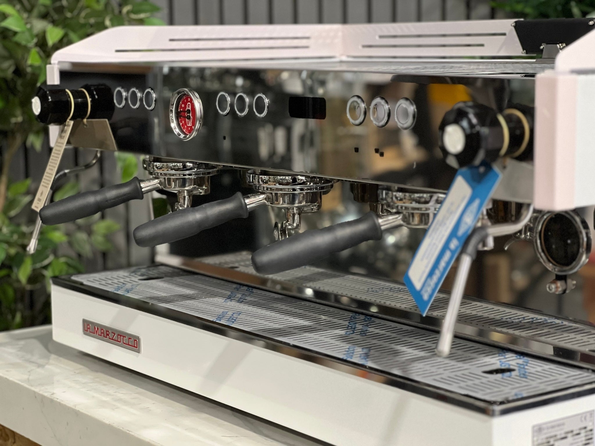 La Marzocco Linea PB 3 Group Coffee Machine White & Pink - New espresso machine close-up angled view, highlighting polished group heads, steam wands, cup tray, and control buttons, placed on a marble bench with greenery behind.