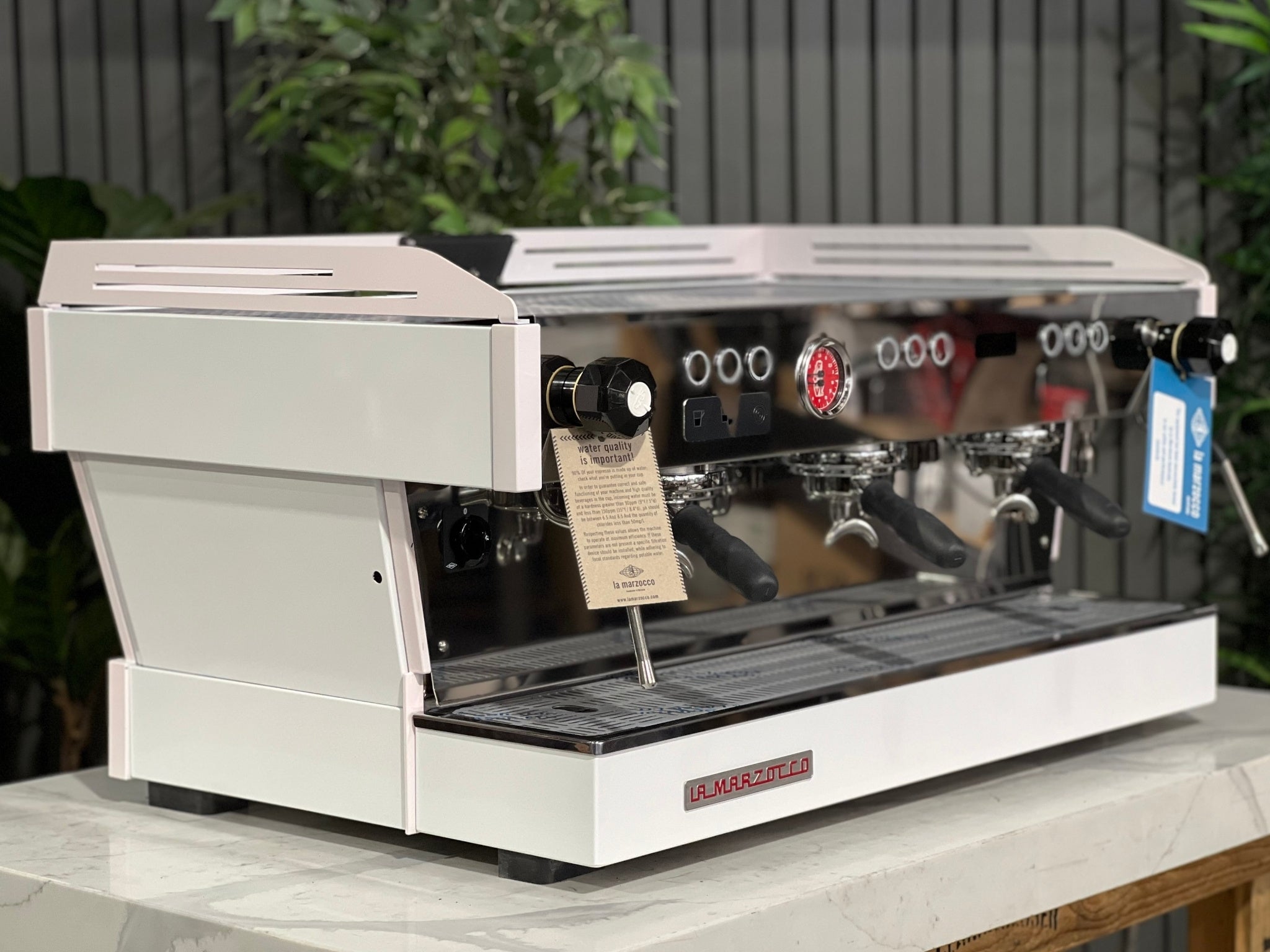 La Marzocco Linea PB 3 Group Coffee Machine White & Pink - New espresso machine angled view, showing three polished group heads, dual steam wands, cup tray, and digital display, placed on a marble bench with greenery in the background.
