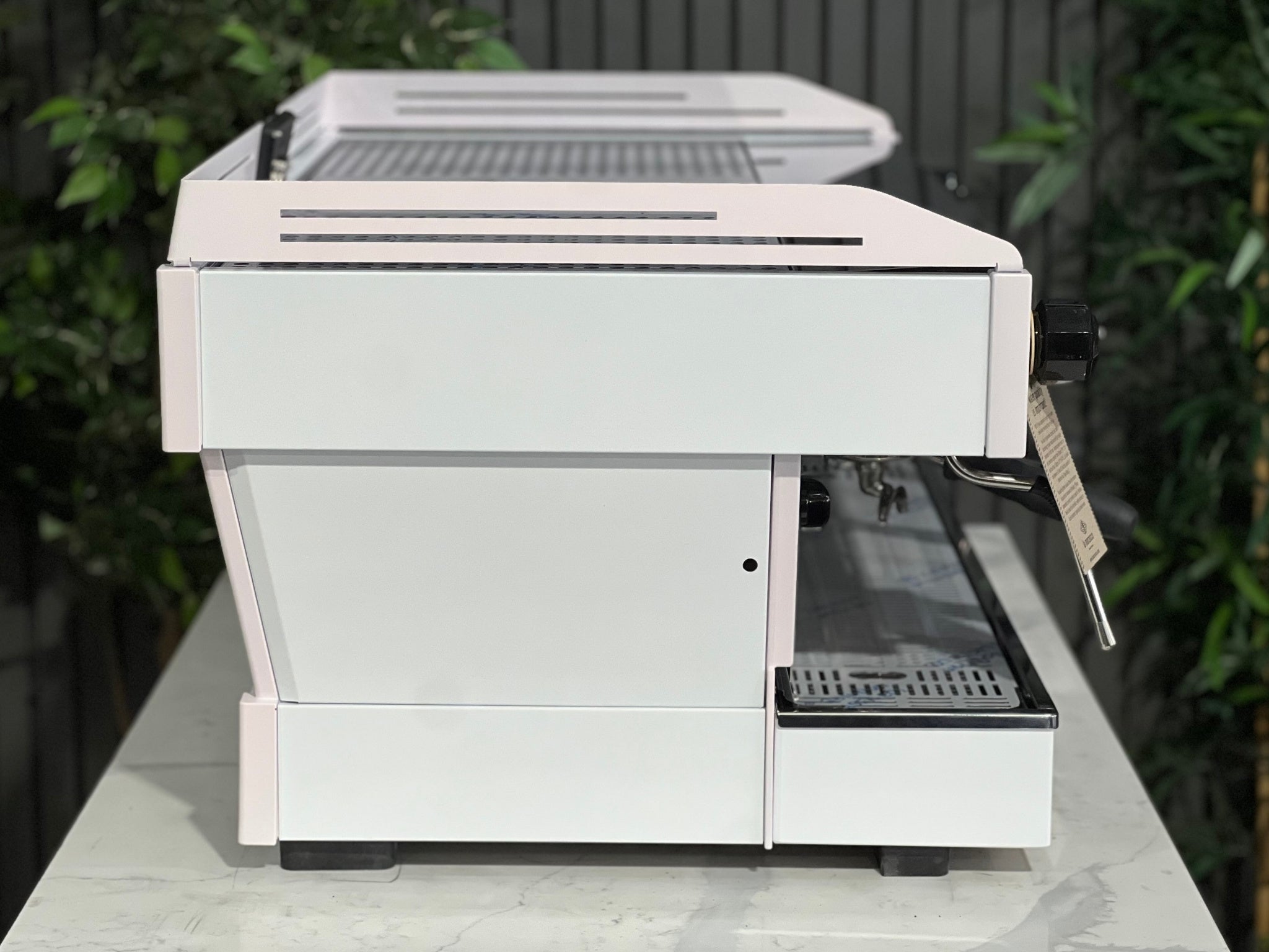 La Marzocco Linea PB 3 Group Coffee Machine White & Pink - New side view, showing the machine’s smooth custom finish, side panel design, and stainless steel frame, positioned on a marble bench with greenery behind.