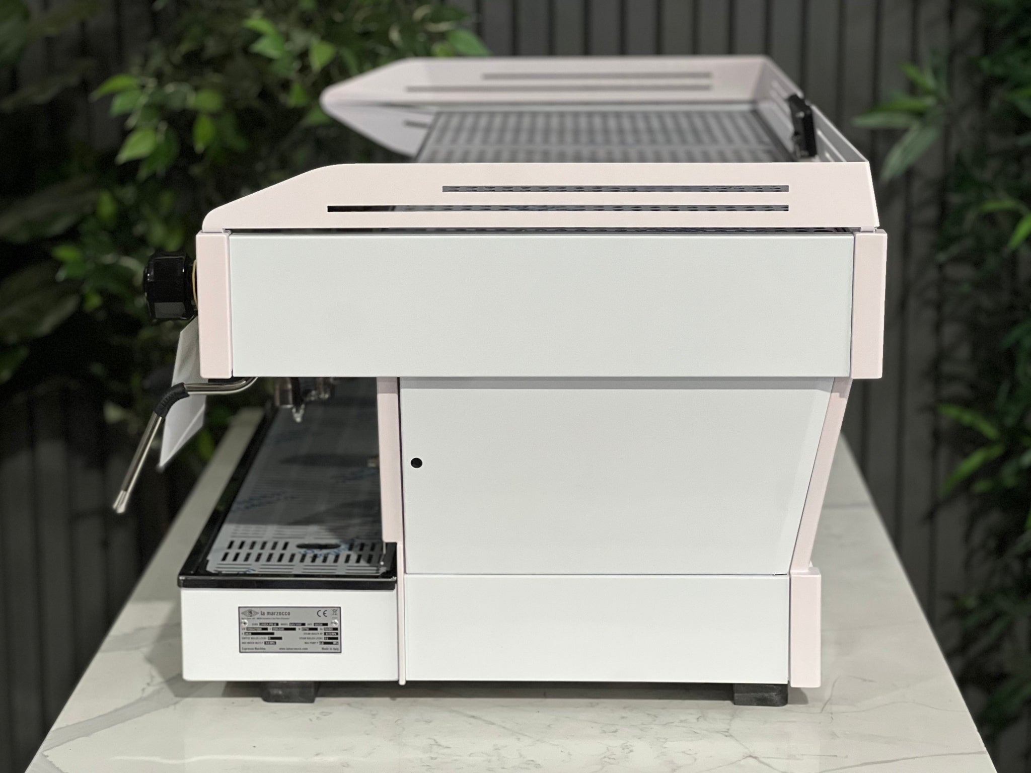 La Marzocco Linea PB 3 Group Coffee Machine White & Pink - New side view, showing the machine’s smooth custom finish, side panel design, and stainless steel frame, positioned on a marble bench with greenery behind.