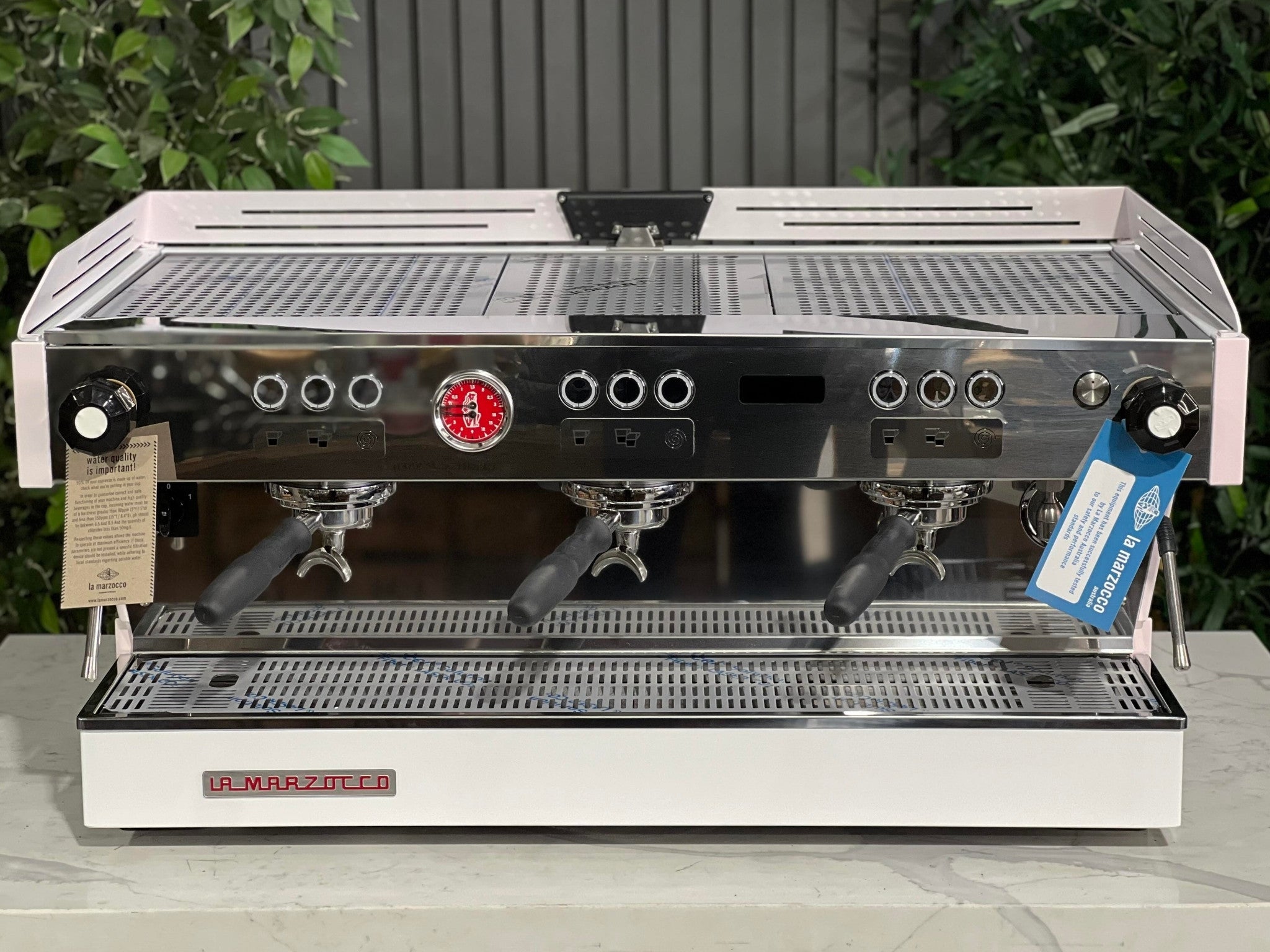 La Marzocco Linea PB 3 Group Coffee Machine White & Pink - New front-on view, highlighting its three group heads, dual steam wands, cup tray, barista lights, and control panel, sitting on a marble bench with greenery behind.