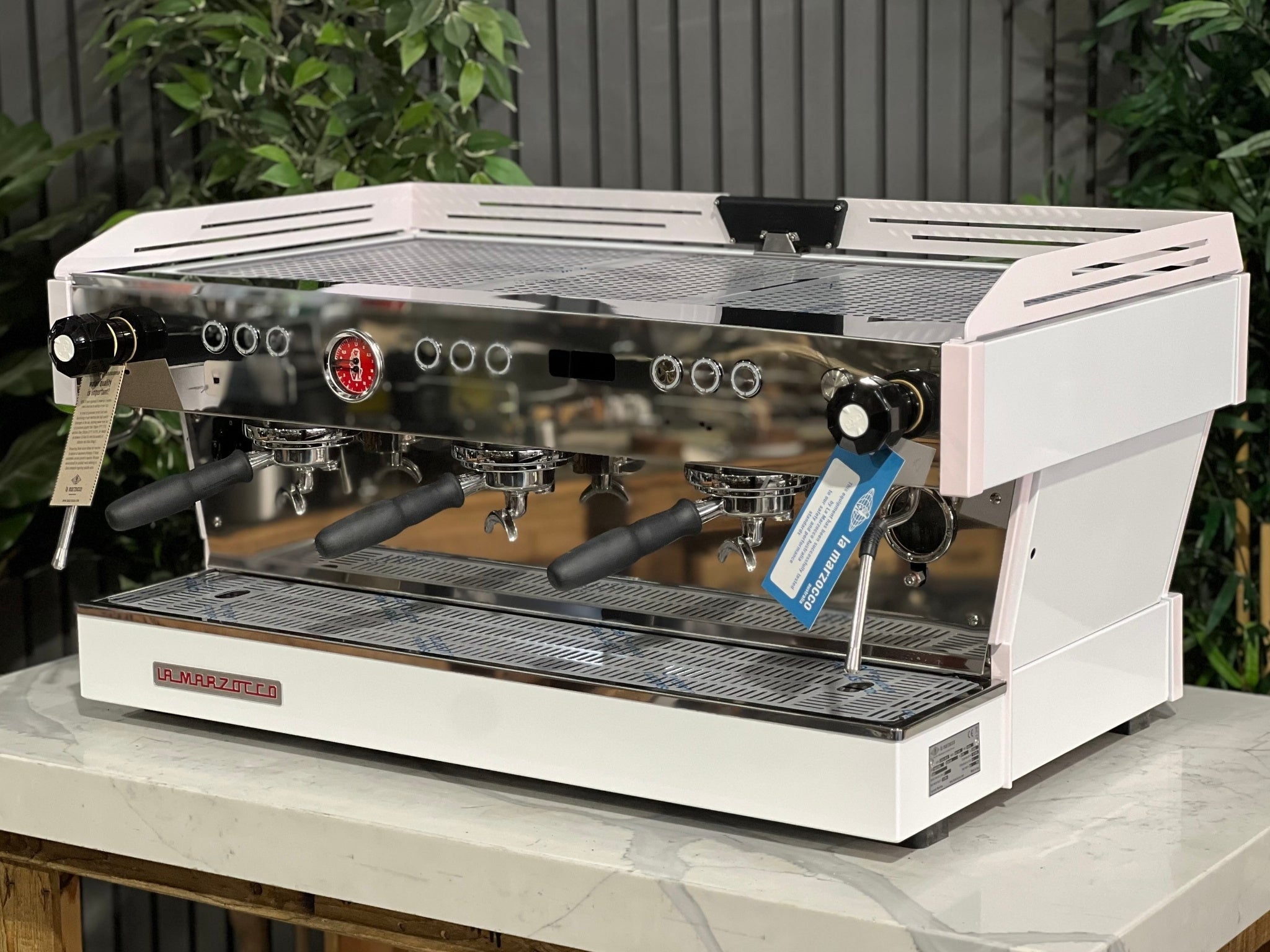 La Marzocco Linea PB 3 Group Coffee Machine White & Pink - New espresso machine angled view, showing three polished group heads, dual steam wands, cup tray, and digital display, placed on a marble bench with greenery in the background.