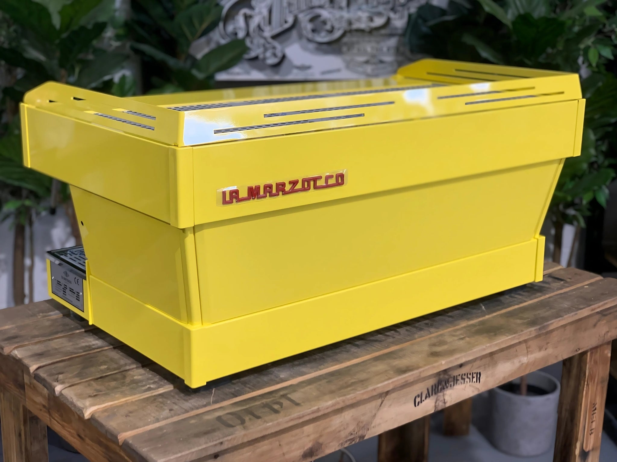 La Marzocco Linea PB 3 Group Coffee Machine Yellow – New espresso coffee machine angled rear view on wooden bench with greenery, showing yellow body, stainless trim, and machine profile.
