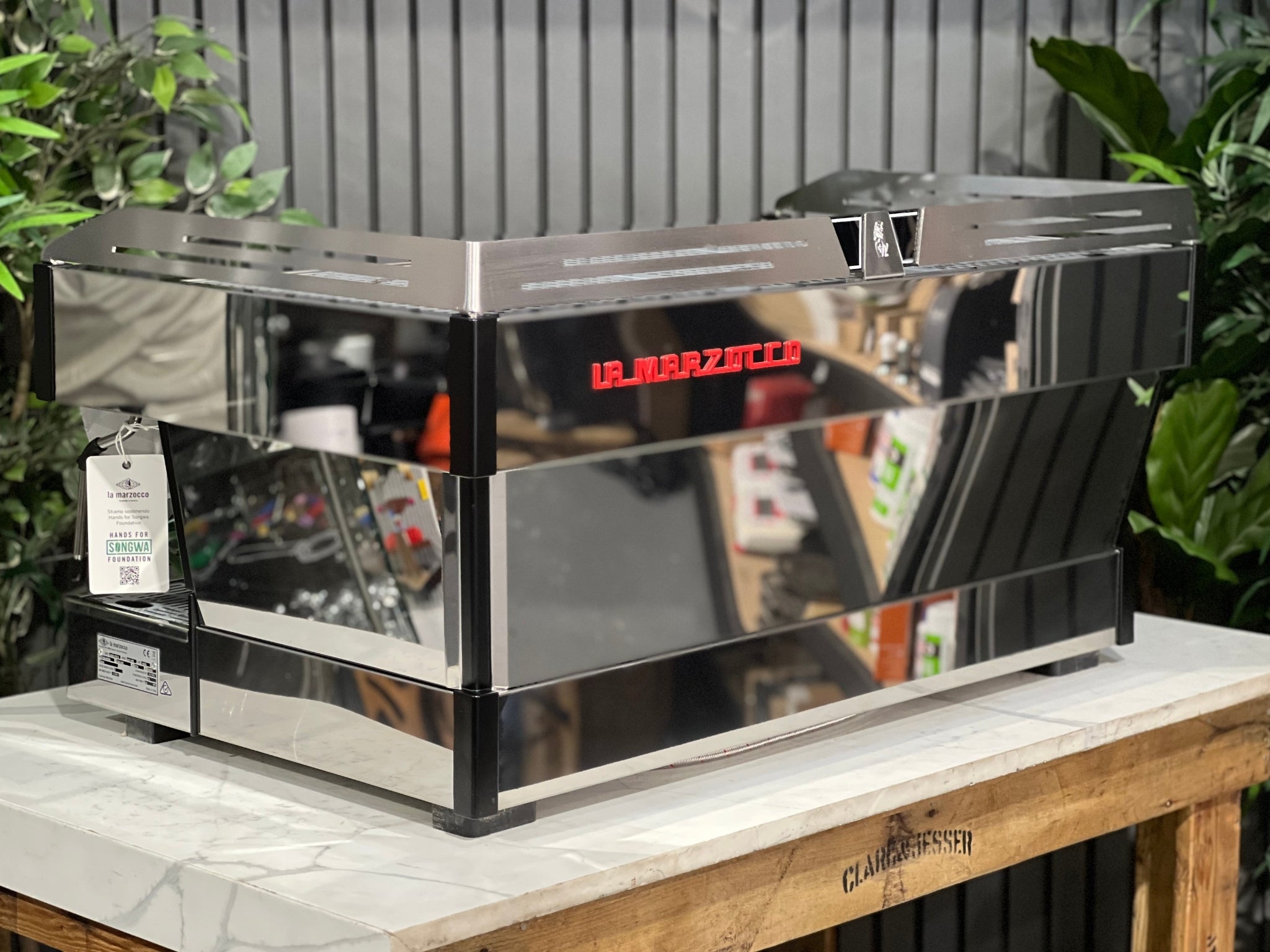 La Marzocco Linea PB ABR 3 Group Coffee Machine Stainless - New, angled back view highlighting stainless steel body, red La Marzocco logo, and side panel details, on a marble bench with greenery in the background.