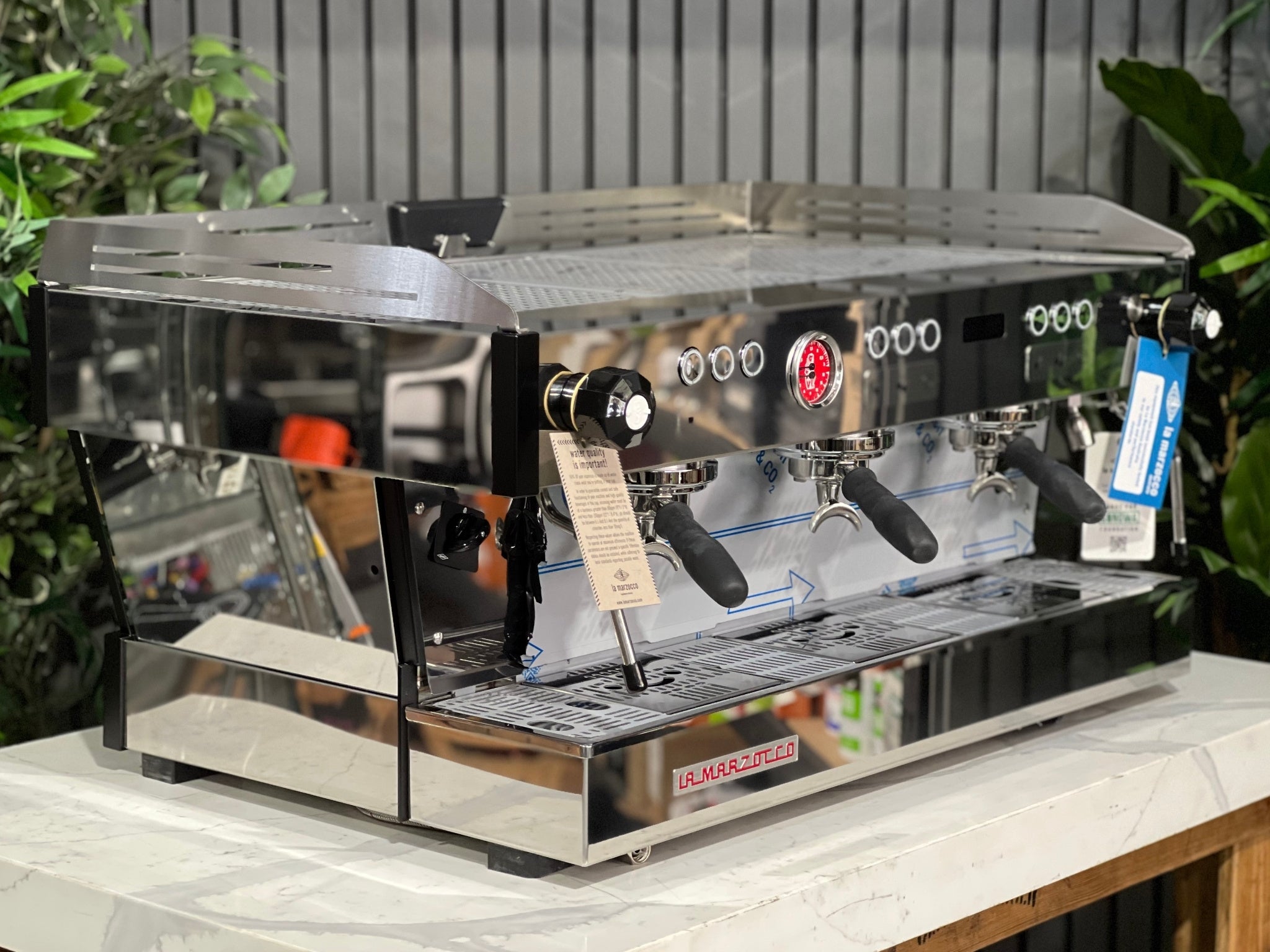 La Marzocco Linea PB ABR 3 Group Coffee Machine Stainless - New, angled view showing stainless steel body, three group heads, dual steam wands, integrated ABR drip tray, digital display, and barista lights, on a marble bench with greenery in the background.