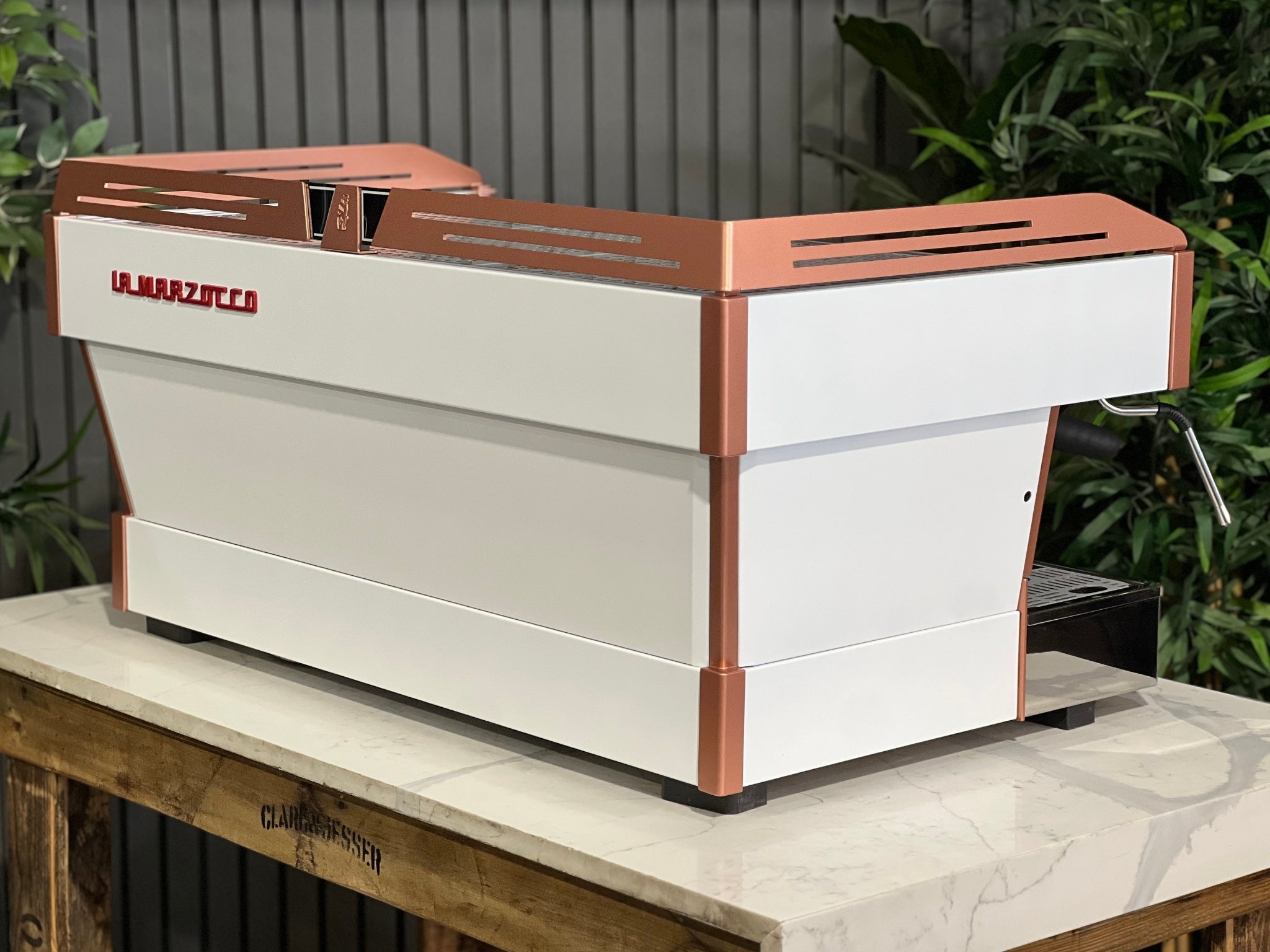 La Marzocco Linea PB ABR 3 Group Coffee Machine White & Copper - New angled back view showing piping, stainless steel rear panels, red La Marzocco logo, and dual boiler system, positioned on a marble bench with greenery behind.