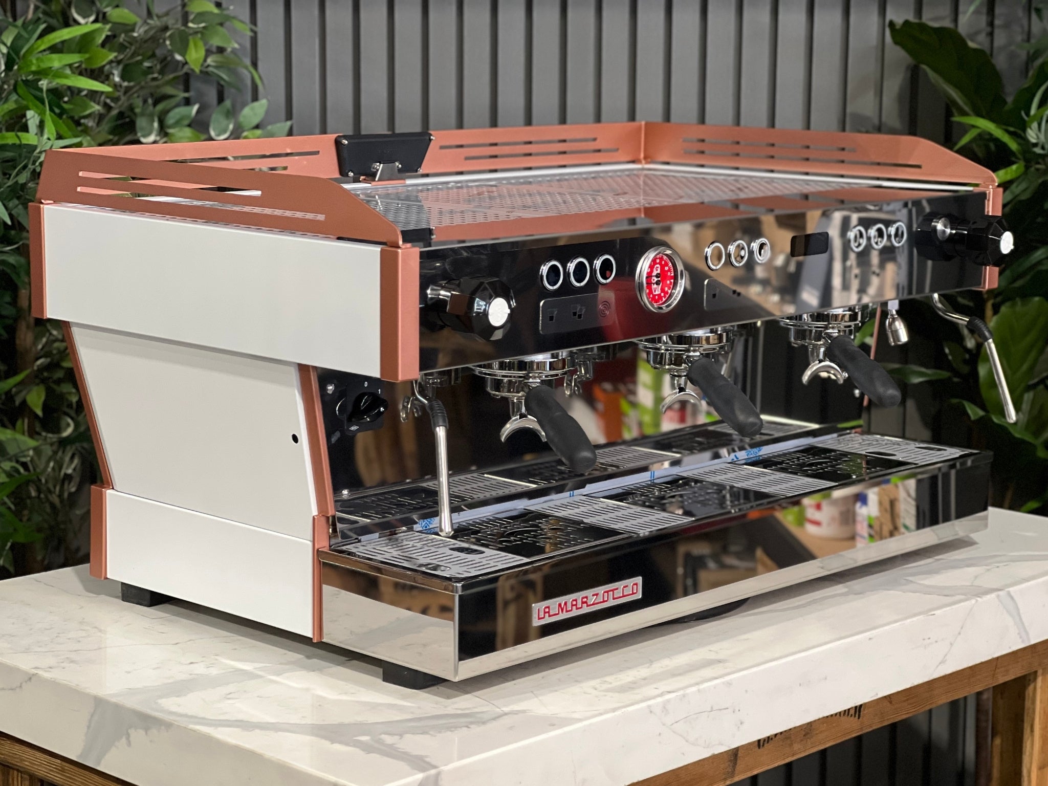 La Marzocco Linea PB ABR 3 Group Coffee Machine White & Copper - New angled view showing its white body with copper accents, dual boiler system, three saturated group heads, dual steam wands, integrated ABR drip tray, digital LED display, and barista lights on a marble bench with greenery in the background.