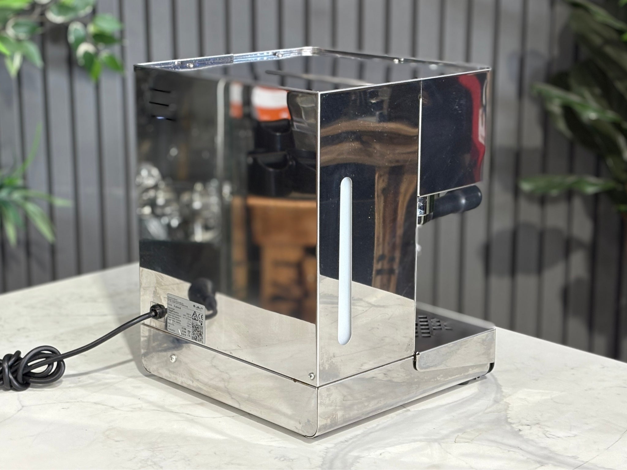 A Lelit Diana PL60 1 Group Coffee Machine with Stainless Steel finish sitting on a marble bench. There is greenery in the background. The image is taken from behind the machine and the machine is sitting on an angle so that you can see the back of the machine and the side. 