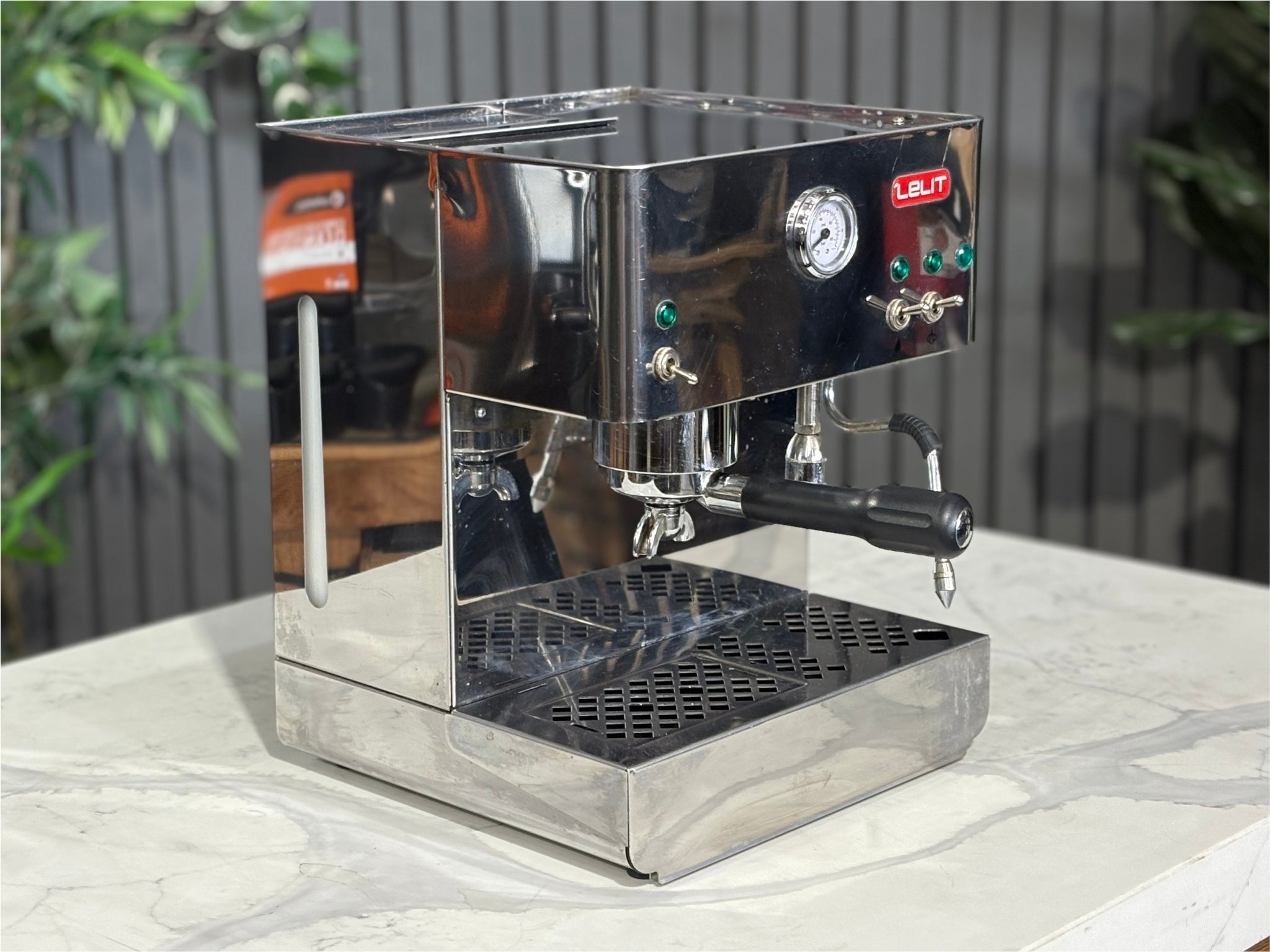 A Lelit Diana PL60 1 Group Coffee Machine with Stainless Steel finish  is sitting on a marble bench. There is greenery in the background. The image is taken from the front of the machine and the machine is sitting on an angle so that you can see the front of the machine and the side. 