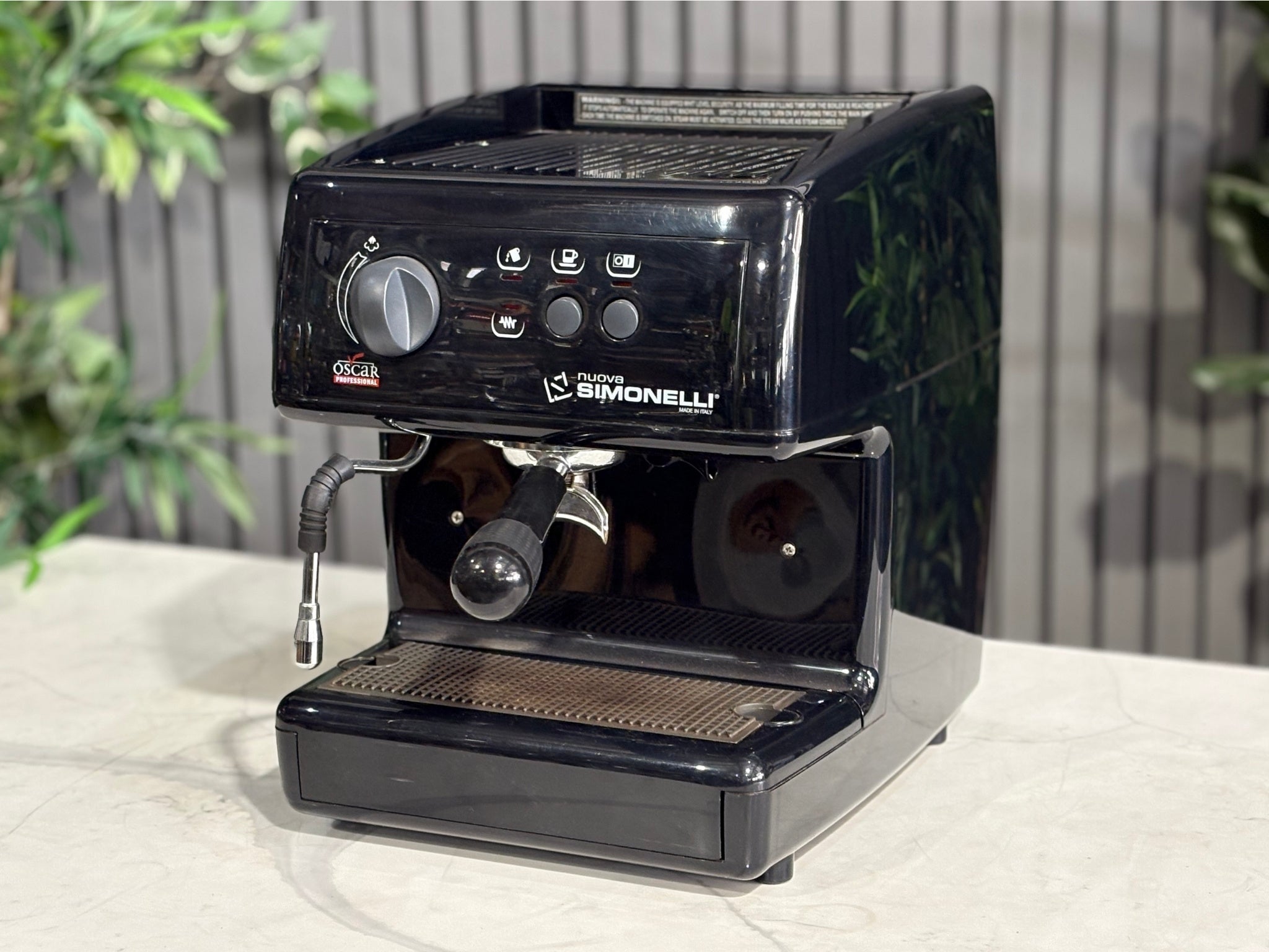 Nuova Simonelli Oscar Professional 1 Group Coffee Machine Black - Used espresso machine displayed at an angle on a marble bench with greenery in the background. Visible features include the button-operated group head, steam wand, and black compact body.
