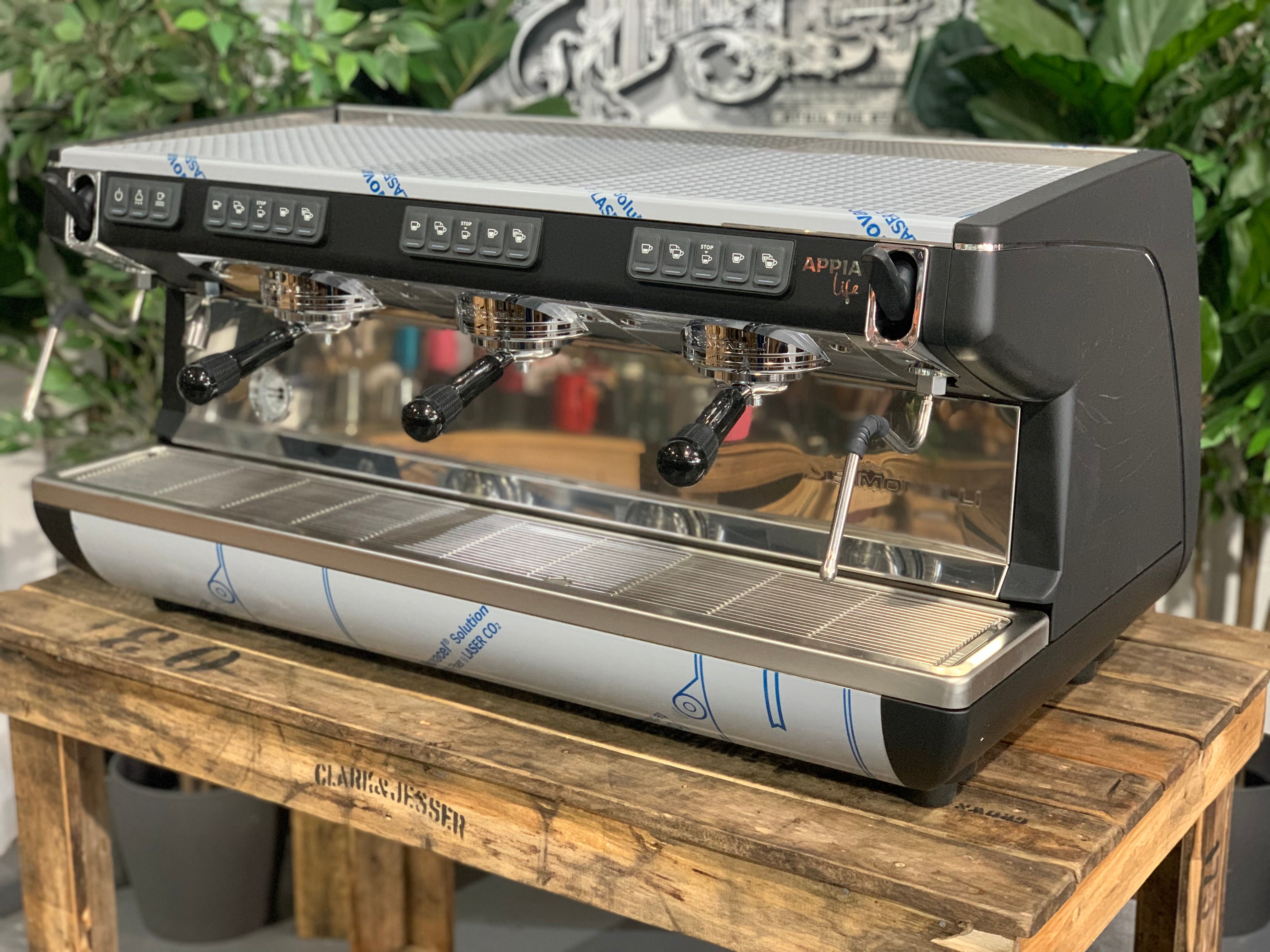 Nuova Simonelli Appia Life Timer 3 Group Coffee Machine Black on a wooden surface with plants in the background.