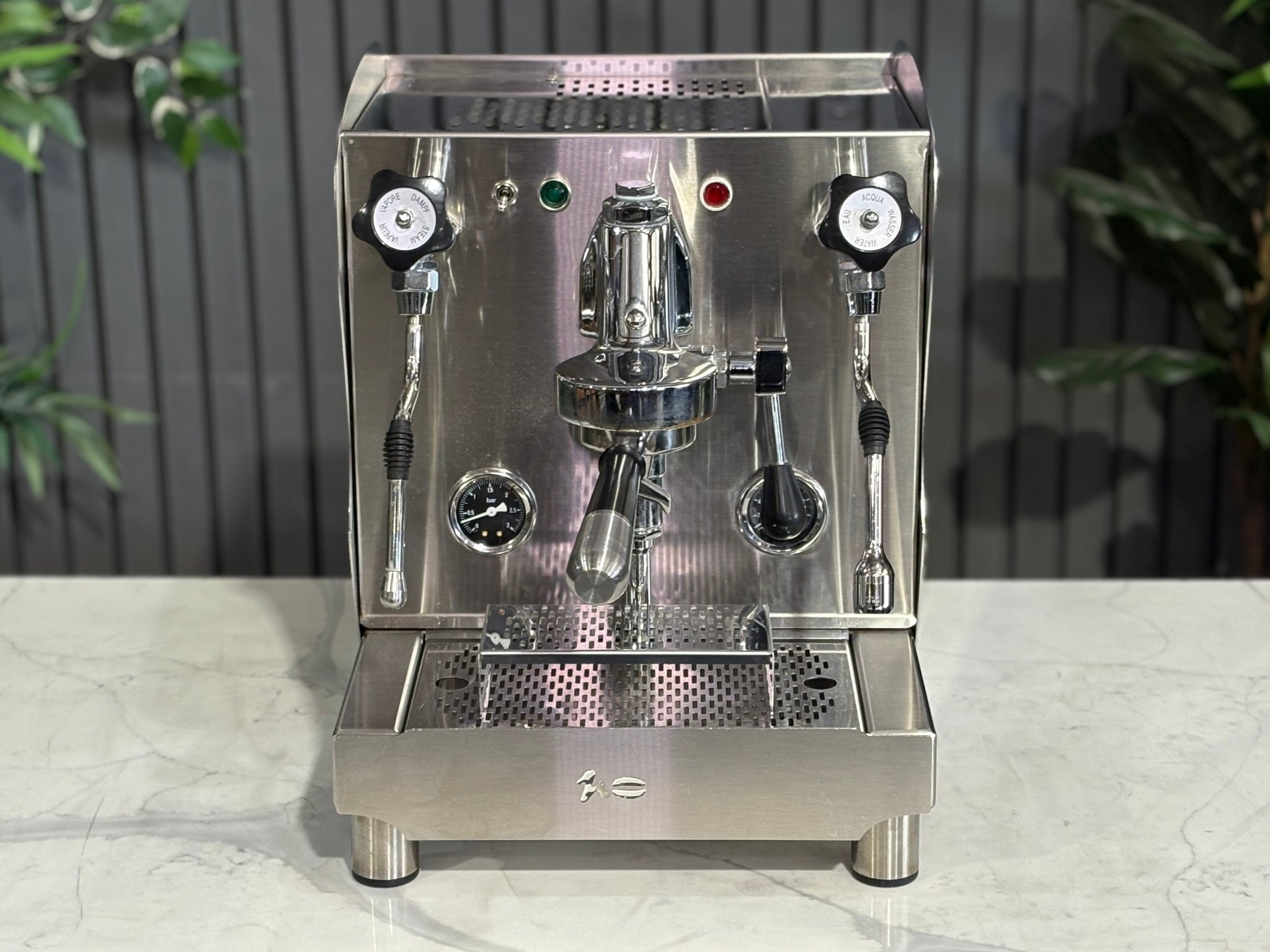 A Orchestrale Nota 1 Group Coffee Machine with Stainless Steel finish sitting on a marble bench. There is greenery in the background. The photo is taken from a high front angle, clearly showcasing both the front panel and the top of the machine.