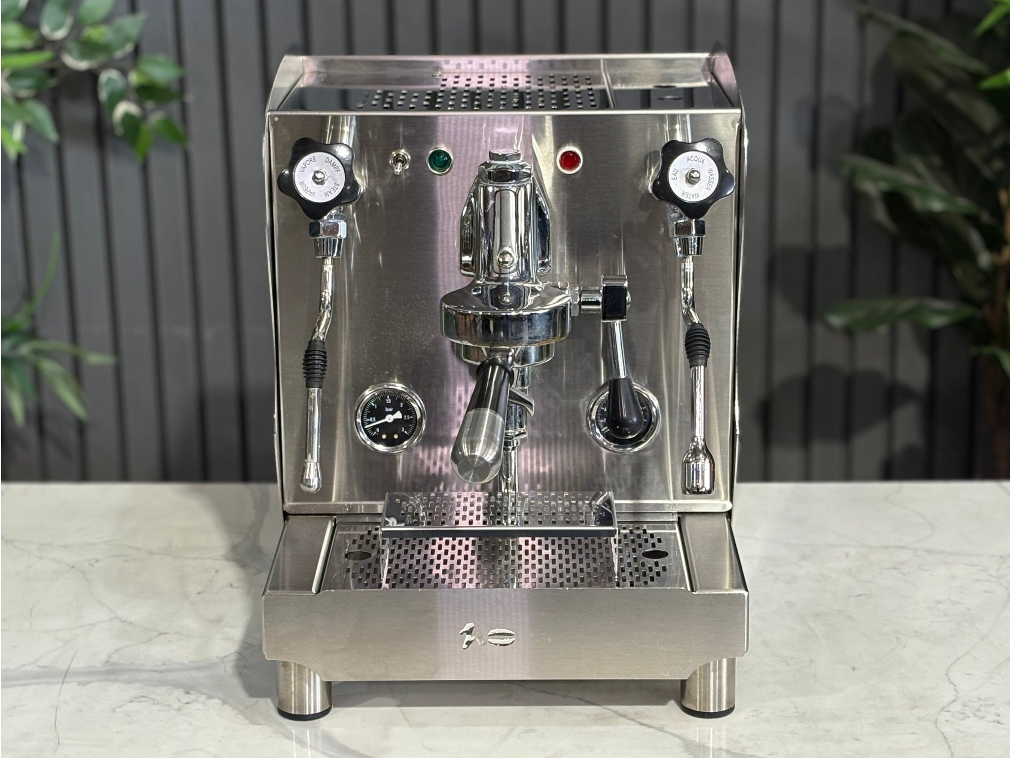 A Orchestrale Nota 1 Group Coffee Machine with Stainless Steel finish  sitting on a marble bench. There is greenery in the background. The photo is taken from a high front angle, clearly showcasing both the front panel and the top of the machine.