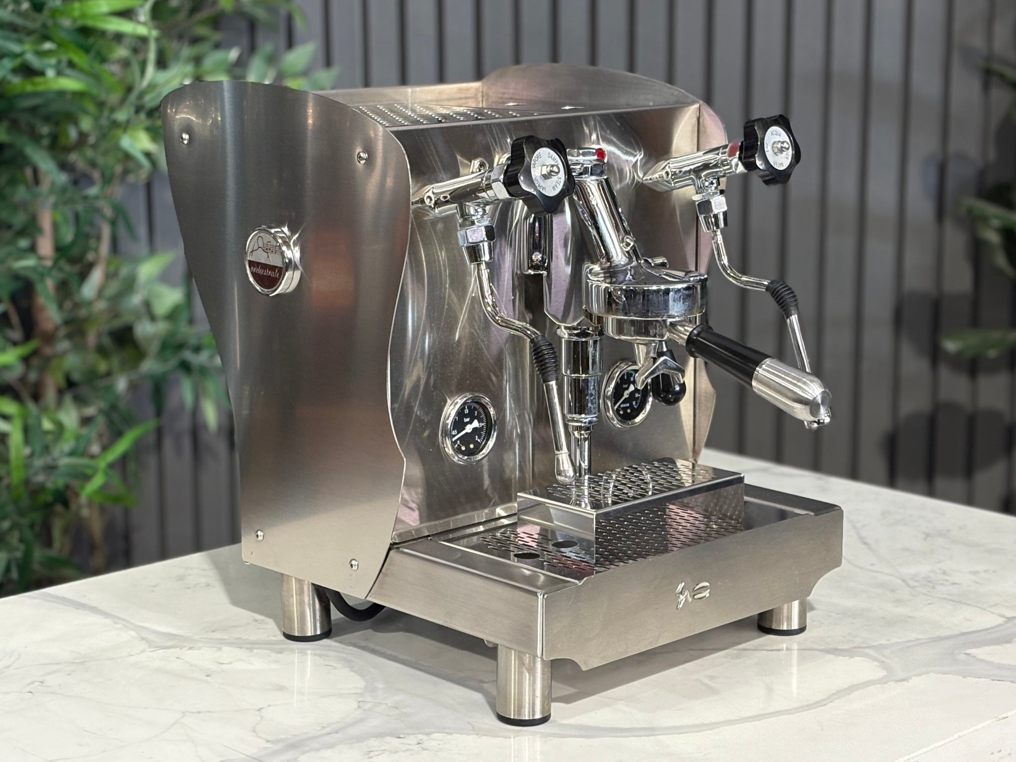 A Orchestrale Nota 1 Group Coffee Machine with Stainless Steel finish  is sitting on a marble bench. There is greenery in the background. The image is taken from the front of the machine and the machine is sitting on an angle so that you can see the front of the machine and the side. 