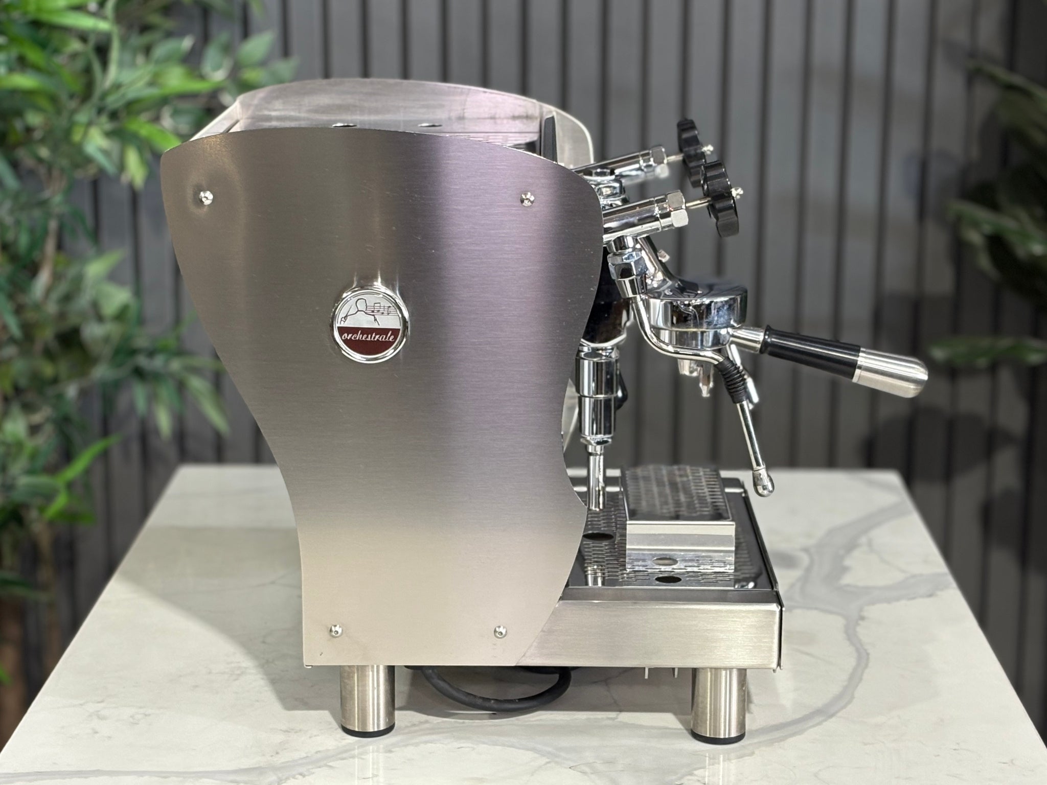 A Orchestrale Nota 1 Group Coffee Machine with Stainless Steel finish  sitting on a marble bench. There is greenery in the background. The image is taken from the side of the machine.