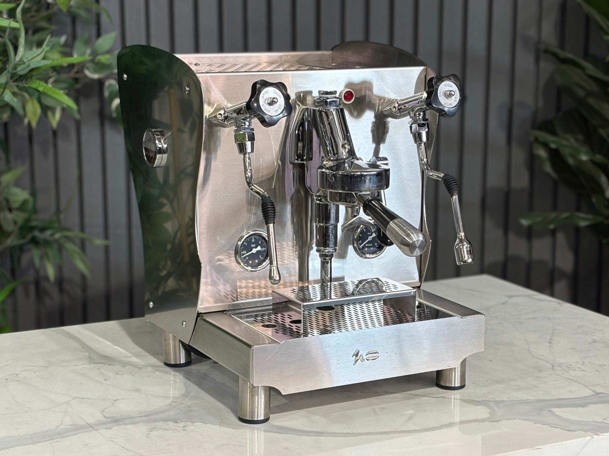 A Orchestrale Nota 1 Group Coffee Machine with Stainless Steel finish l is sitting on a marble bench. There is greenery in the background. The image is taken from the front of the machine and the machine is sitting on an angle so that you can see the front of the machine and the side. 