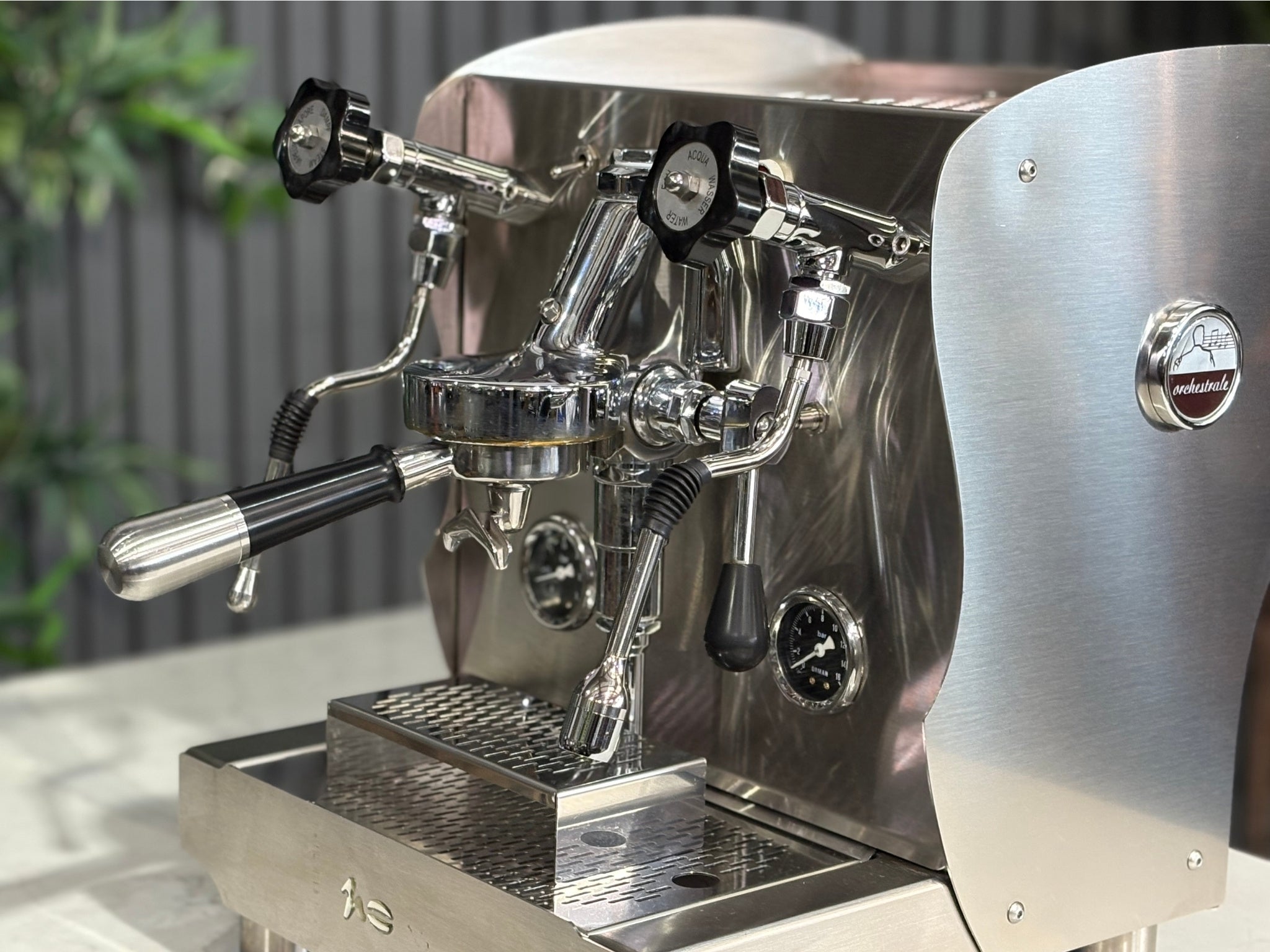 A Orchestrale Nota 1 Group Coffee Machine with Stainless Steel finish  is sitting on a marble bench. There is greenery in the background. The image is a close up image taken from the front of the machine and the machine is sitting on an angle so that you can see the front of the machine, the controls including rotary knobs and, group handle.