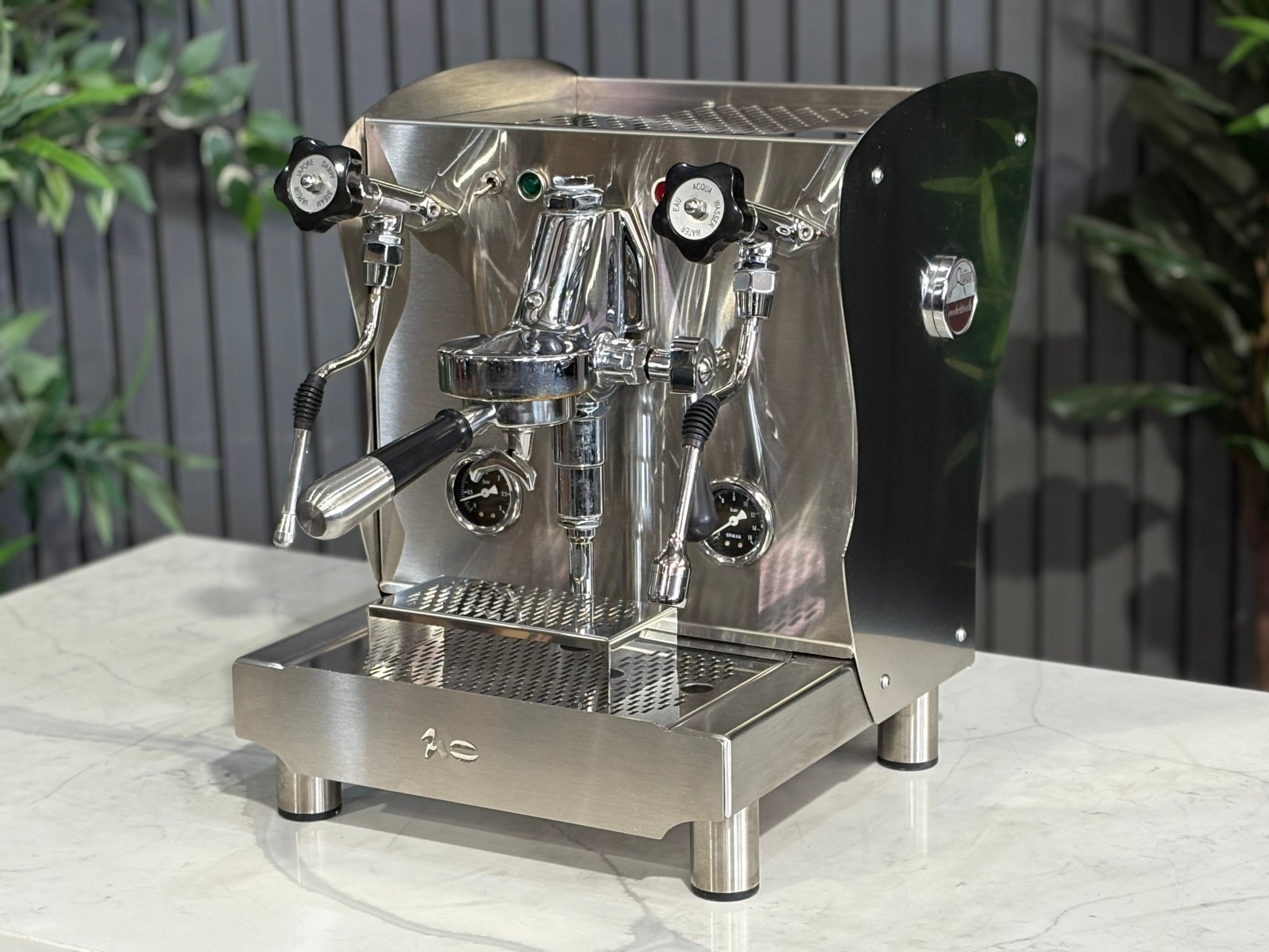 A Orchestrale Nota 1 Group Coffee Machine with Stainless Steel finish is sitting on a marble bench. There is greenery in the background. The image is taken from the front of the machine and the machine is sitting on an angle so that you can see the front of the machine and the side. 