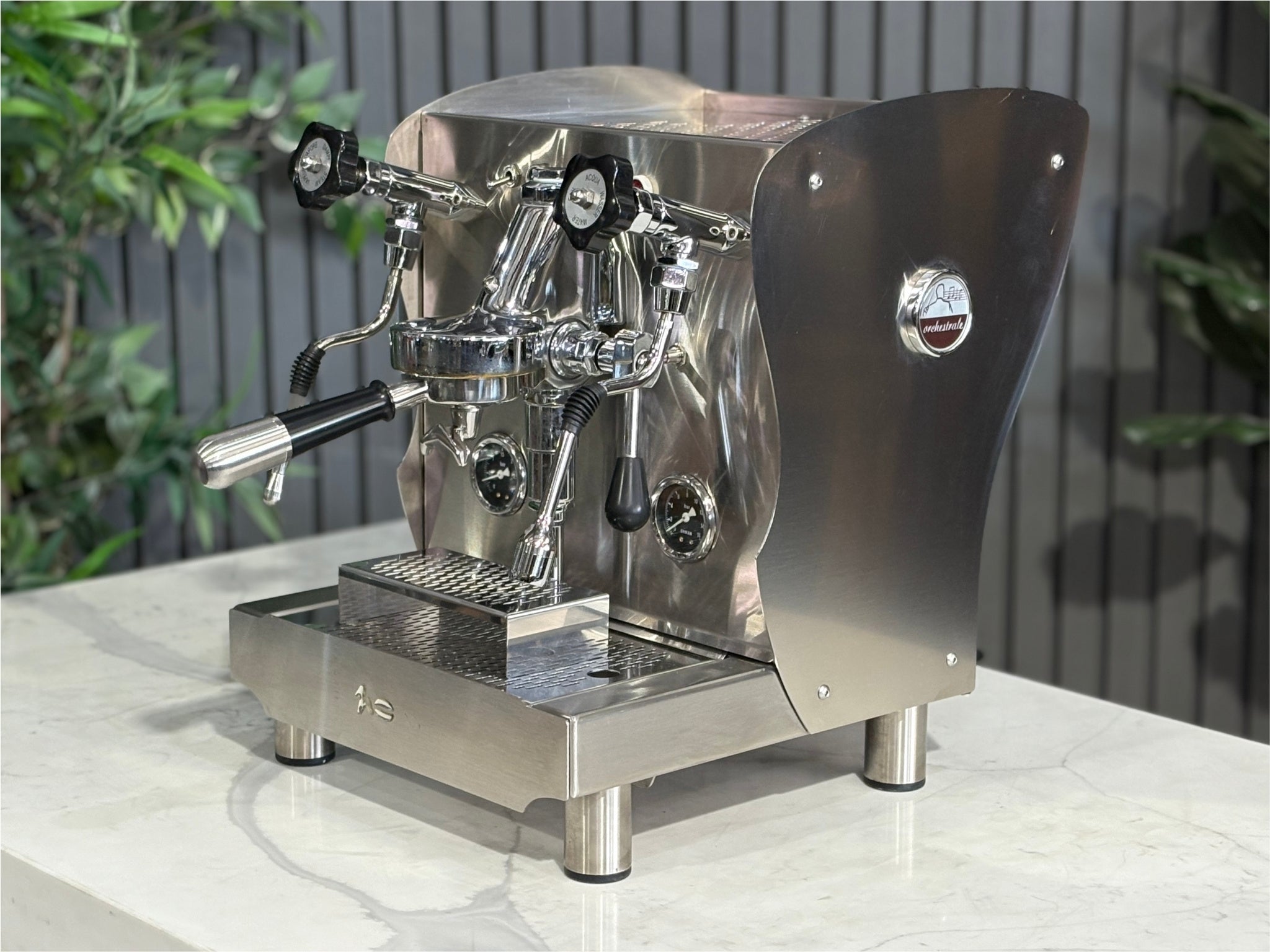 A Orchestrale Nota 1 Group Coffee Machine with Stainless Steel finish is sitting on a marble bench. There is greenery in the background. The image is taken from the front of the machine and the machine is sitting on an angle so that you can see the front of the machine and the side. 