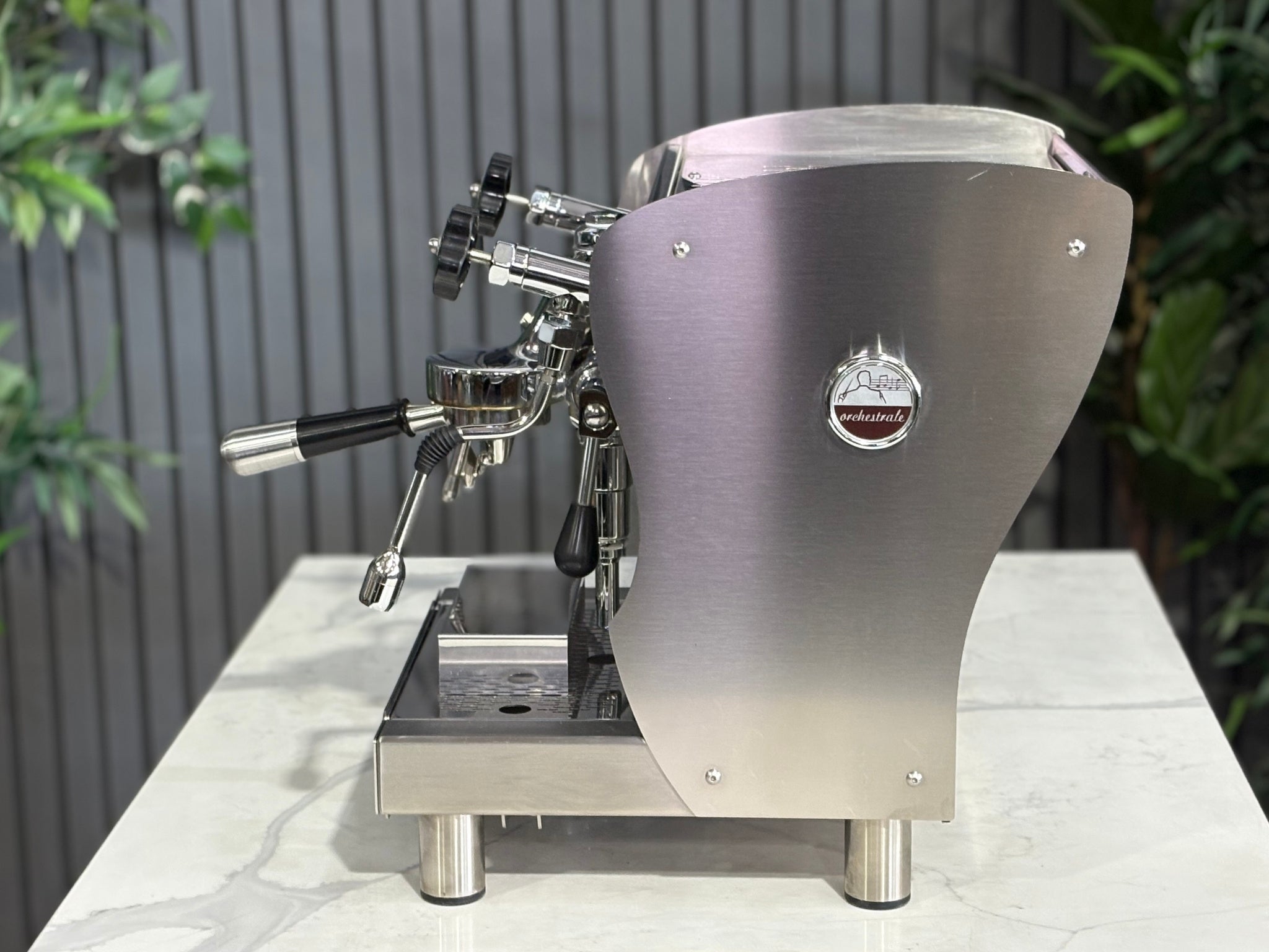 A Orchestrale Nota 1 Group Coffee Machine with Stainless Steel finish  sitting on a marble bench. There is greenery in the background. The image is taken from the side of the machine.