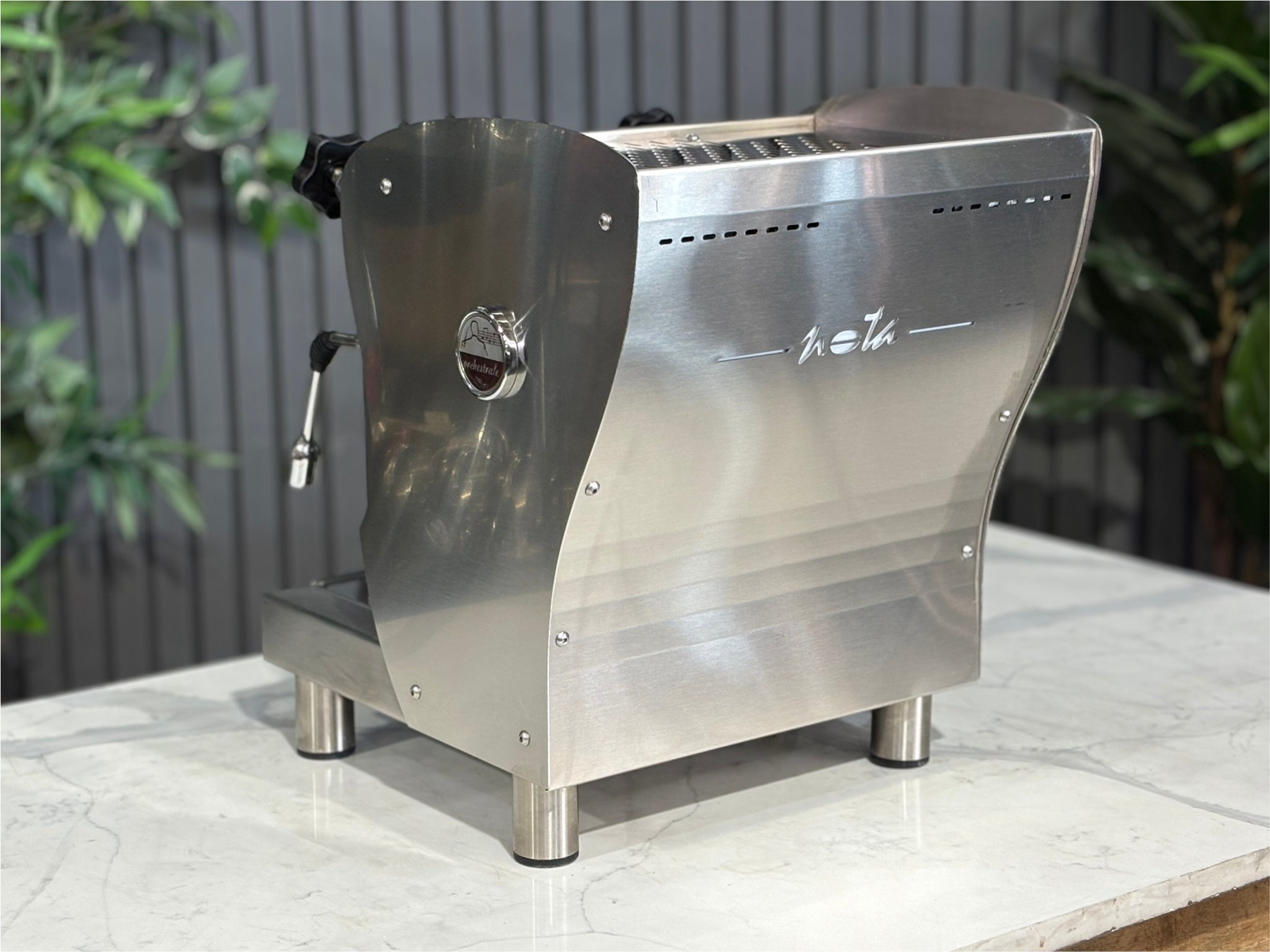 A Orchestrale Nota 1 Group Coffee Machine with Stainless Steel finish sitting on a marble bench. There is greenery in the background. The image is taken from behind the machine and the machine is sitting on an angle so that you can see the back of the machine and the side.