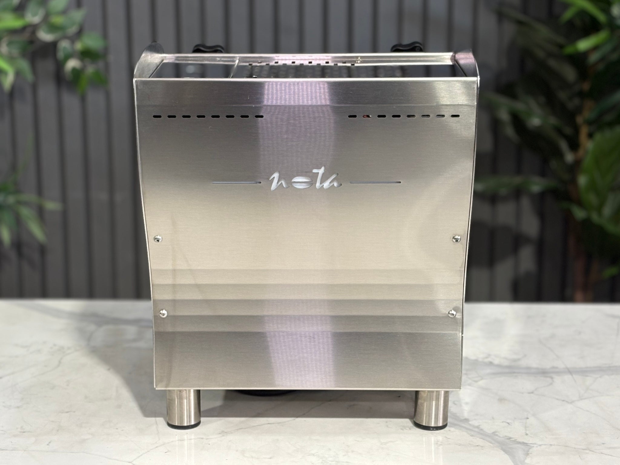 A Orchestrale Nota 1 Group Coffee Machine with Stainless Steel finish is sitting on a marble bench. There is greenery in the background. The image is taken from behind the machine so the back is visible.