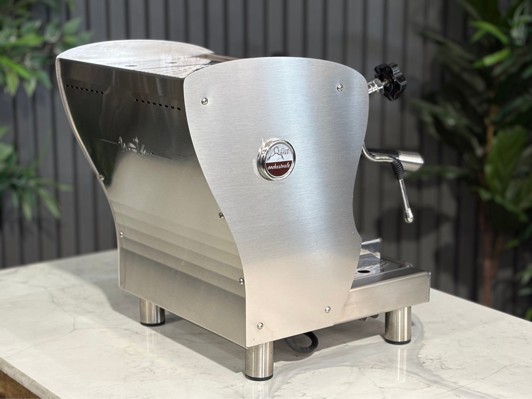 A Orchestrale Nota 1 Group Coffee Machine with Stainless Steel finish  sitting on a marble bench. There is greenery in the background. The image is taken from behind the machine and the machine is sitting on an angle so that you can see the back of the machine and the side.