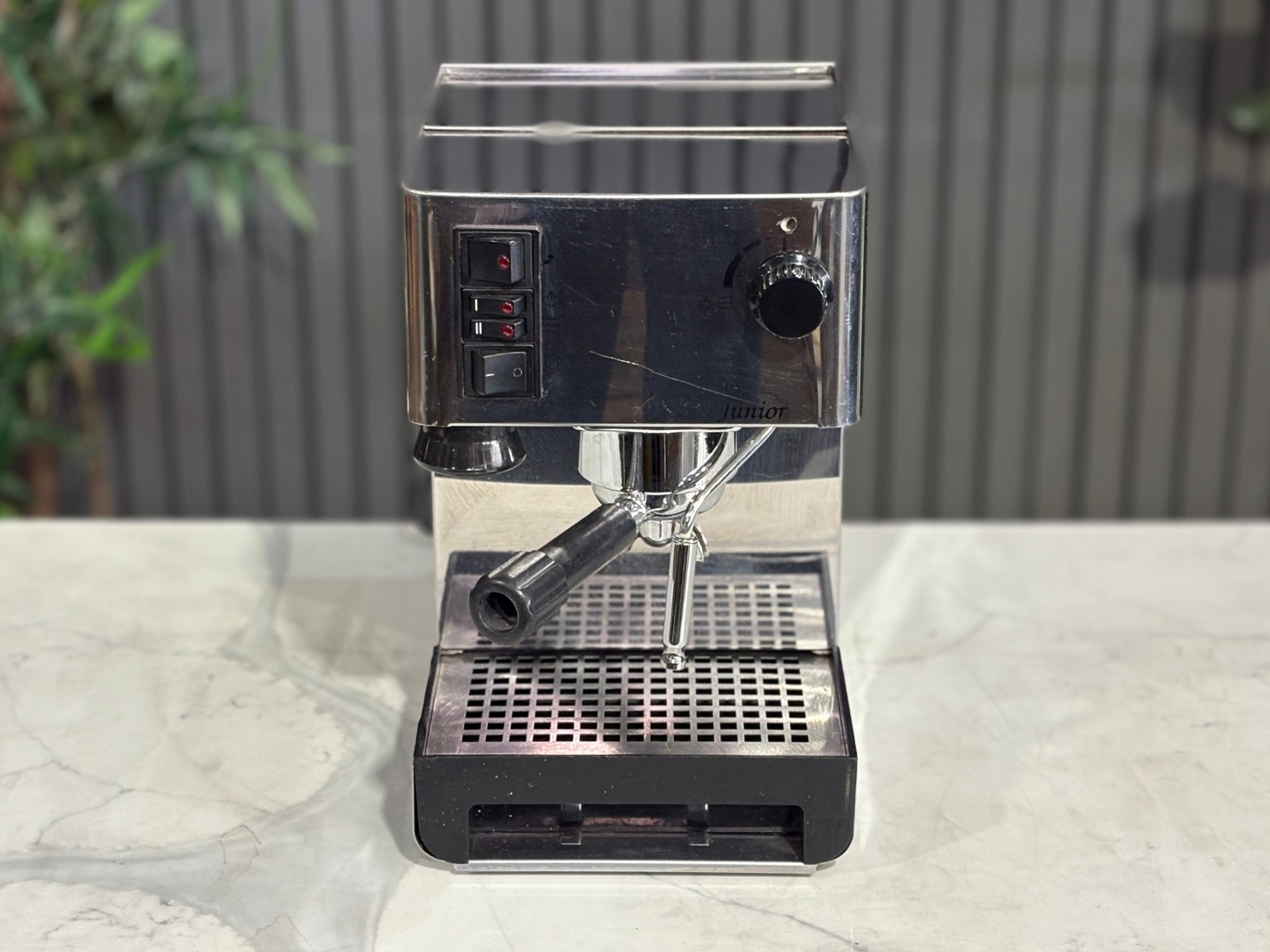A Quaha Lelit Junior Serie II 1 Group Espresso Coffee Machine with Stainless finish sitting on a marble bench. There is greenery in the background. The photo is taken from a high front angle, clearly showcasing both the front panel and the top of the machine.