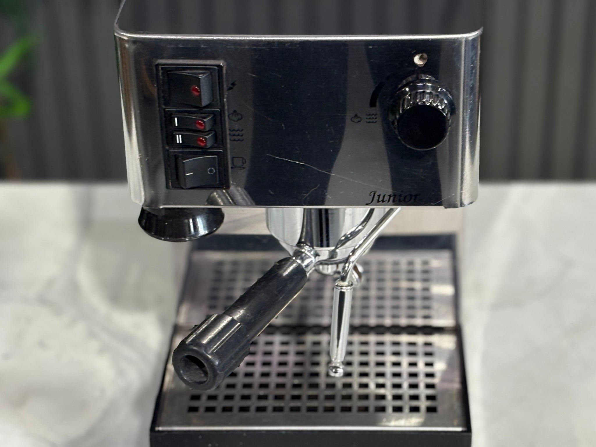A Quaha Lelit Junior Serie II 1 Group Espresso Coffee Machine with Stainless finish is sitting on a marble bench. There is greenery in the background. The image is a close up image taken from the front of the machine so that the controls including flick buttons, group handle and logo is visable.
