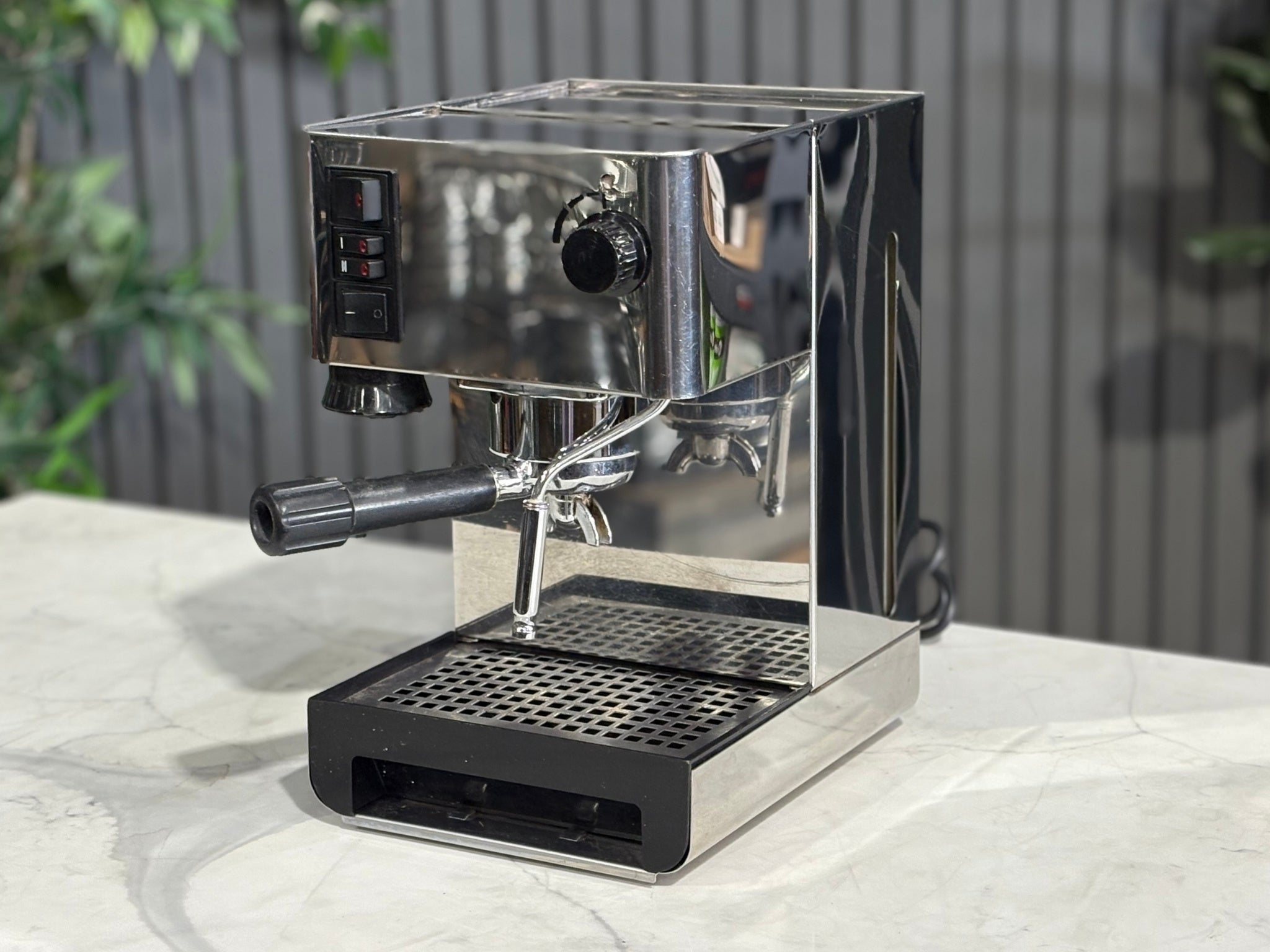 A Quaha Lelit Junior Serie II 1 Group Espresso Coffee Machine with Stainless finish  is sitting on a marble bench. There is greenery in the background. The image is taken from the front of the machine and the machine is sitting on an angle so that you can see the front of the machine and the side. 