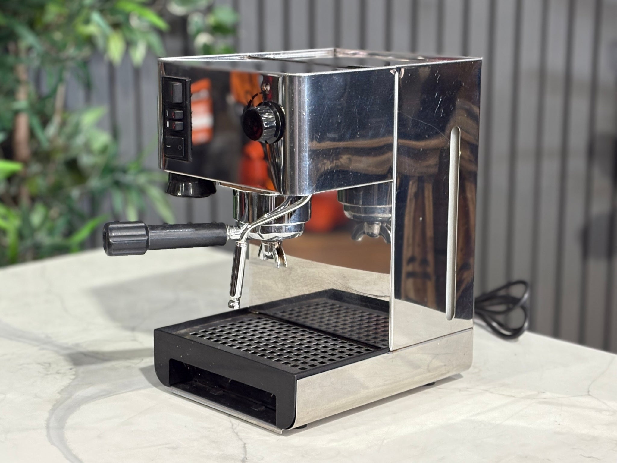 A Quaha Lelit Junior Serie II 1 Group Espresso Coffee Machine with Stainless finish  is sitting on a marble bench. There is greenery in the background. The image is taken from the front of the machine and the machine is sitting on an angle so that you can see the front of the machine and the side. 