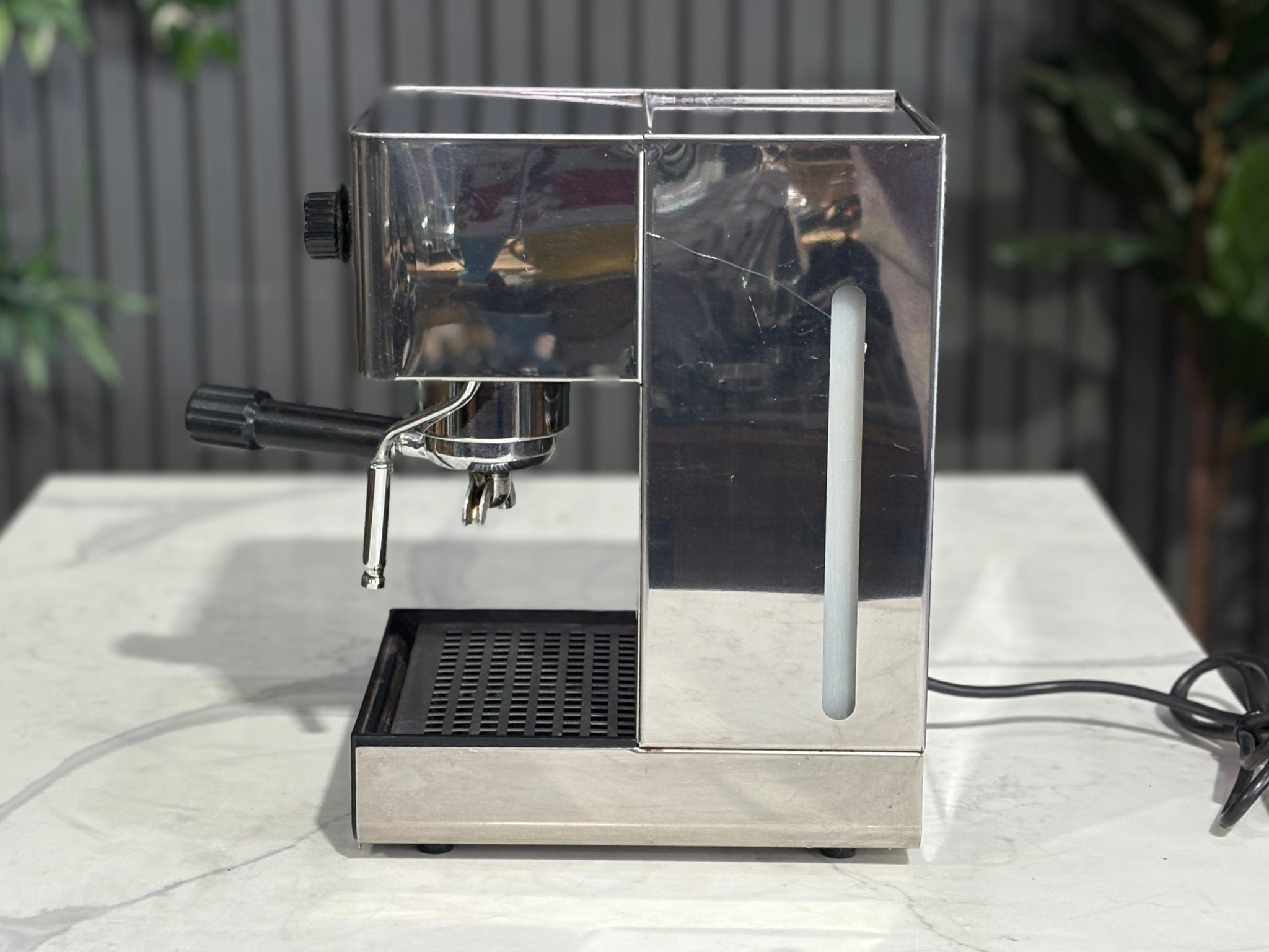 A Quaha Lelit Junior Serie II 1 Group Espresso Coffee Machine with Stainless finish  sitting on a marble bench. There is greenery in the background. The image is taken from the side of the machine.