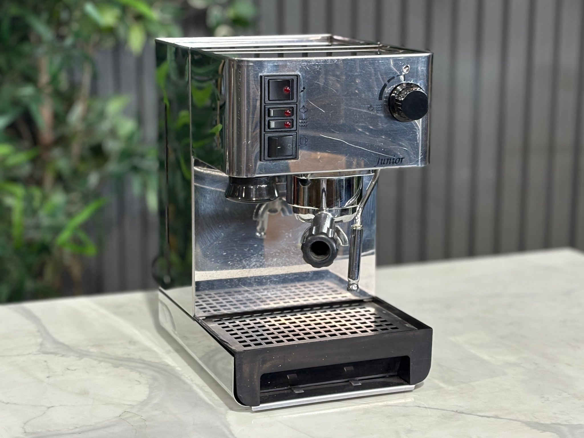 A Quaha Lelit Junior Serie II 1 Group Espresso Coffee Machine with Stainless finish  is sitting on a marble bench. There is greenery in the background. The image is taken from the front of the machine and the machine is sitting on an angle so that you can see the front of the machine and the side. 