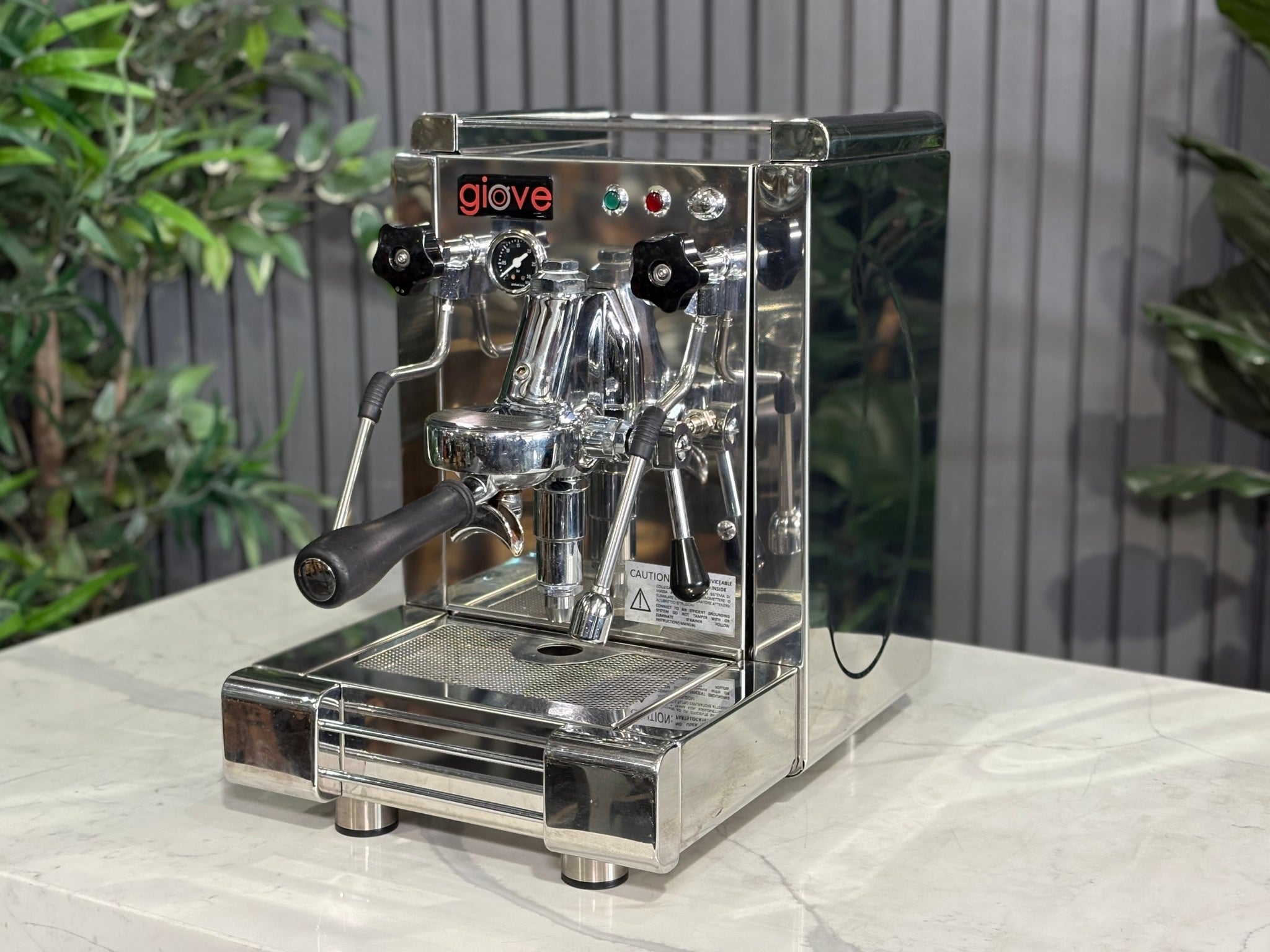 A Royal First Giove 1 Group Espresso Coffee Machine with Stainless finish  is sitting on a marble bench. There is greenery in the background. The image is taken from the front of the machine and the machine is sitting on an angle so that you can see the front of the machine and the side. 