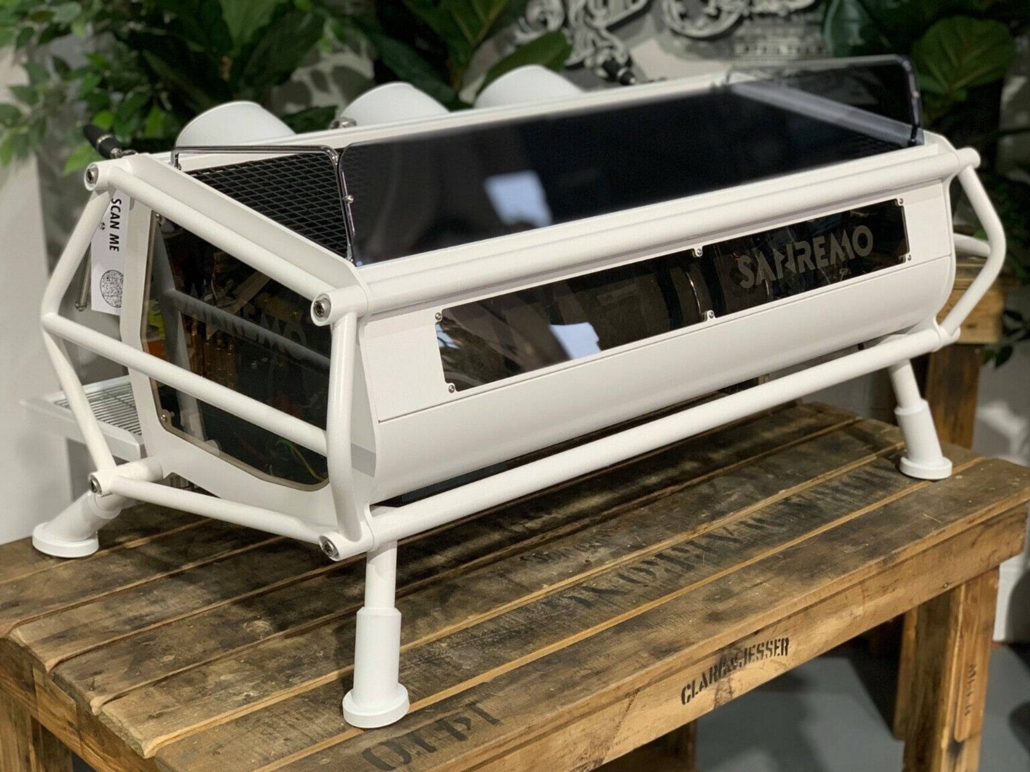 A new San Remo Cafe Racer 3 Group Coffee Machine Naked with white finish sitting on a wooden bench. There is greenery in the background. The image is taken from behind the machine and the machine is sitting on an angle so that you can see the back of the machine and the side.