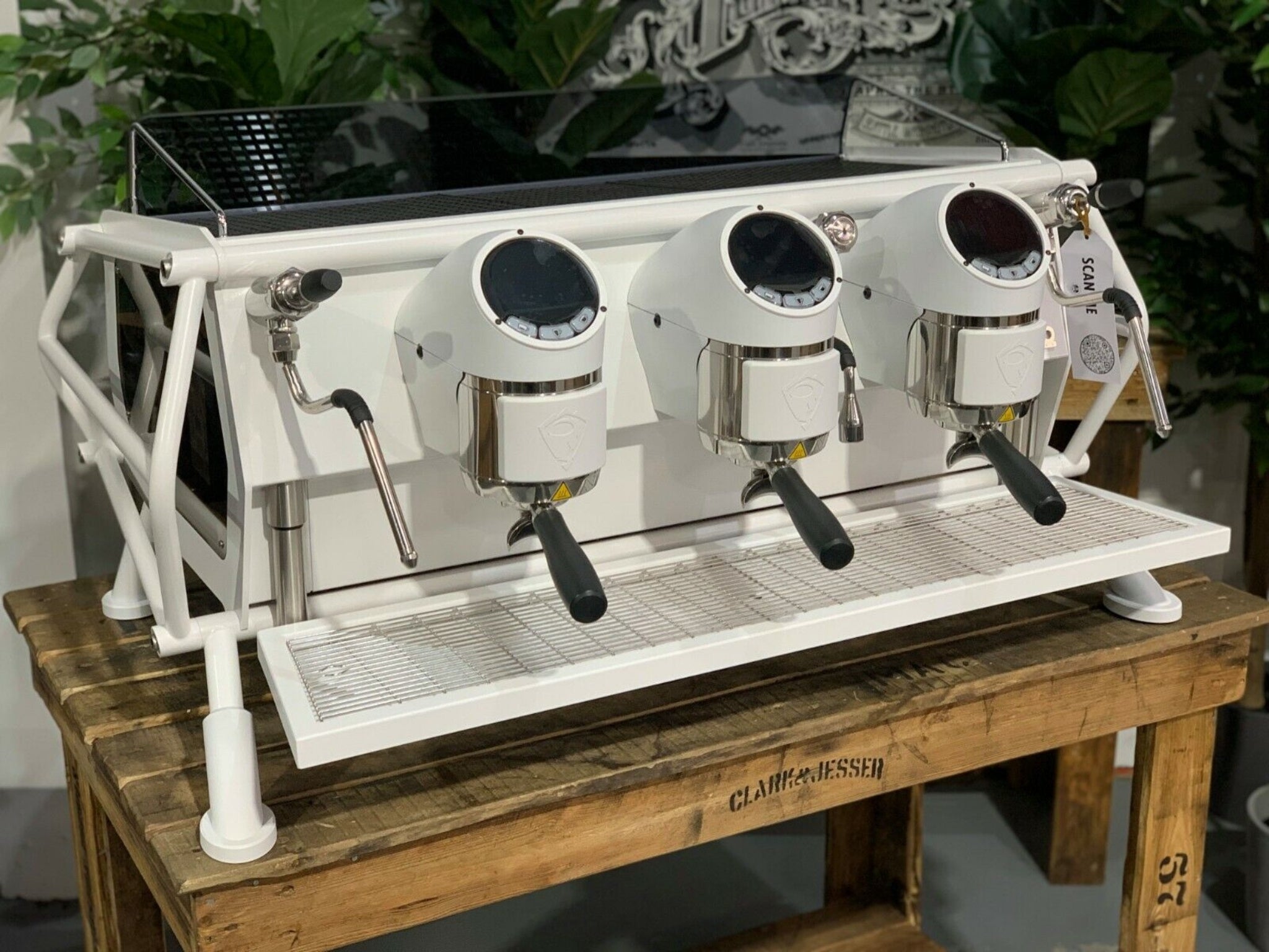 A new San Remo Cafe Racer 3 Group Coffee Machine Naked with white finish
is sitting on a wooden bench. There is greenery in the background. The image is taken from the front of the machine and the machine is sitting on an angle so that you can see the front of the machine and the side.
