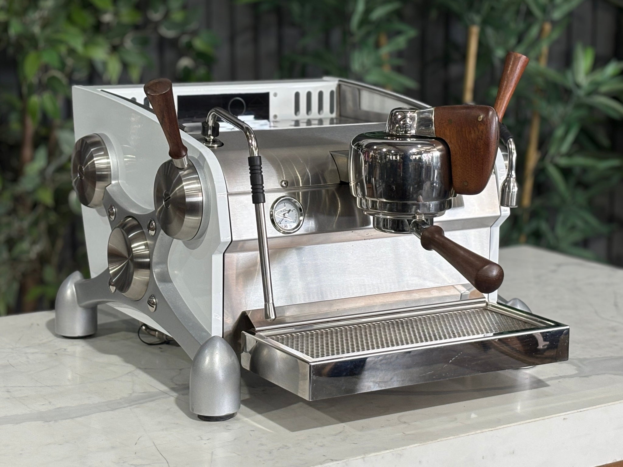 Slayer Espresso 1 Group Coffee Machine White – Used, angled view showing the front and side of the espresso coffee machine with white panels, timber accents, manual paddle, and dual steam wands on a marble bench with greenery in the background.
