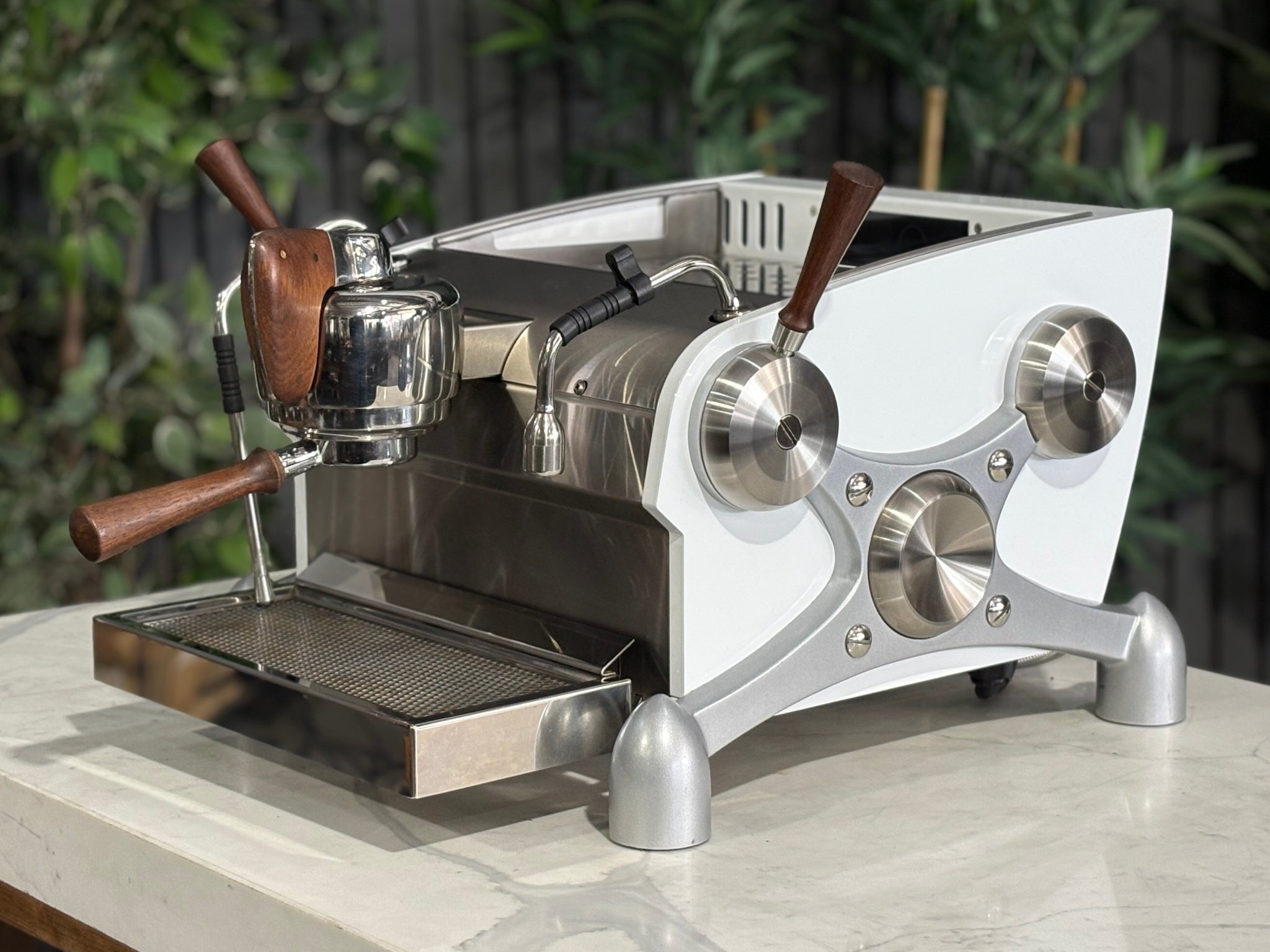Slayer Espresso 1 Group Coffee Machine White – Used, angled view showing the front and side of the espresso coffee machine with white panels, timber accents, manual paddle, and dual steam wands on a marble bench with greenery in the background.