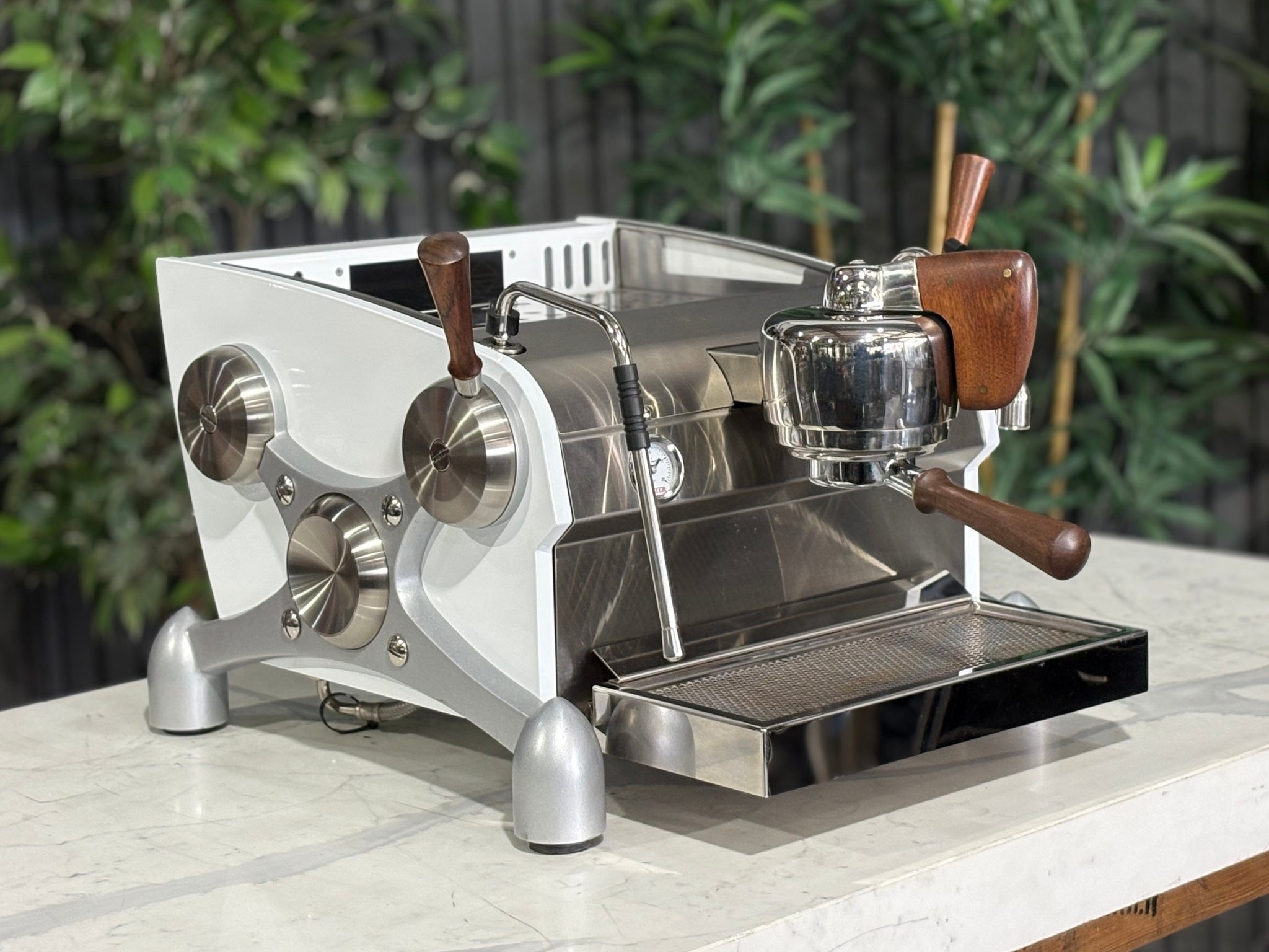 Slayer Espresso 1 Group Coffee Machine White – Used, angled view showing the front and side of the espresso coffee machine with white panels, timber accents, manual paddle, and dual steam wands on a marble bench with greenery in the background.