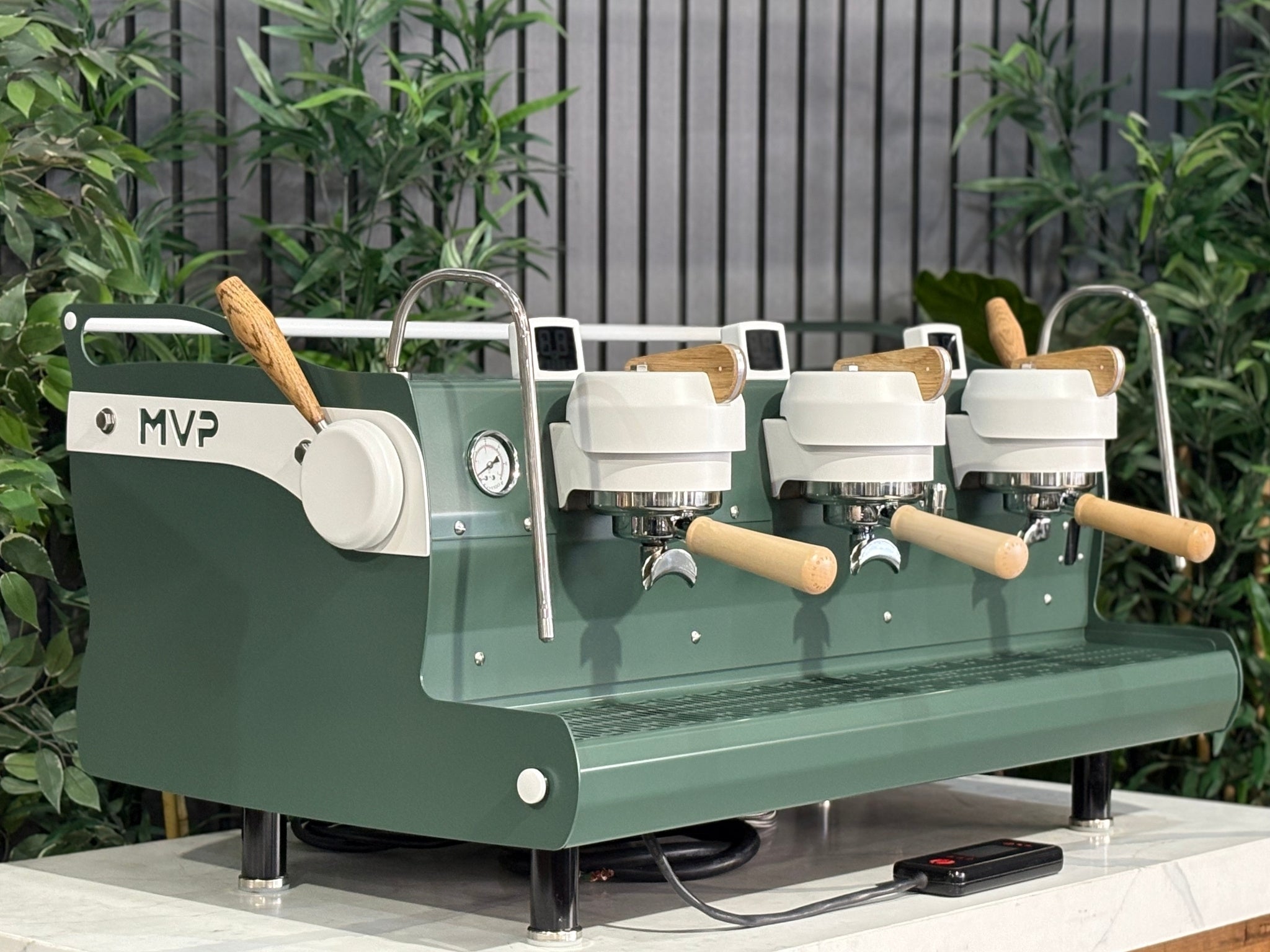 Synesso MVP 3 Group Coffee Machine Green, White & Timber - Used, angled view showing green and white painted body with timber accents, dual group heads, cool touch steam wands, cup tray, and digital shot timers on a marble bench with greenery in the background.