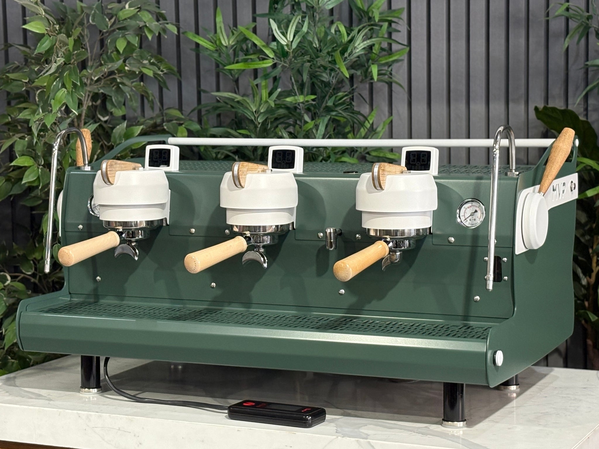 Synesso MVP 3 Group Coffee Machine Green, White & Timber - Used, angled view showing green and white painted body with timber accents, dual group heads, cool touch steam wands, cup tray, and digital shot timers on a marble bench with greenery in the background.