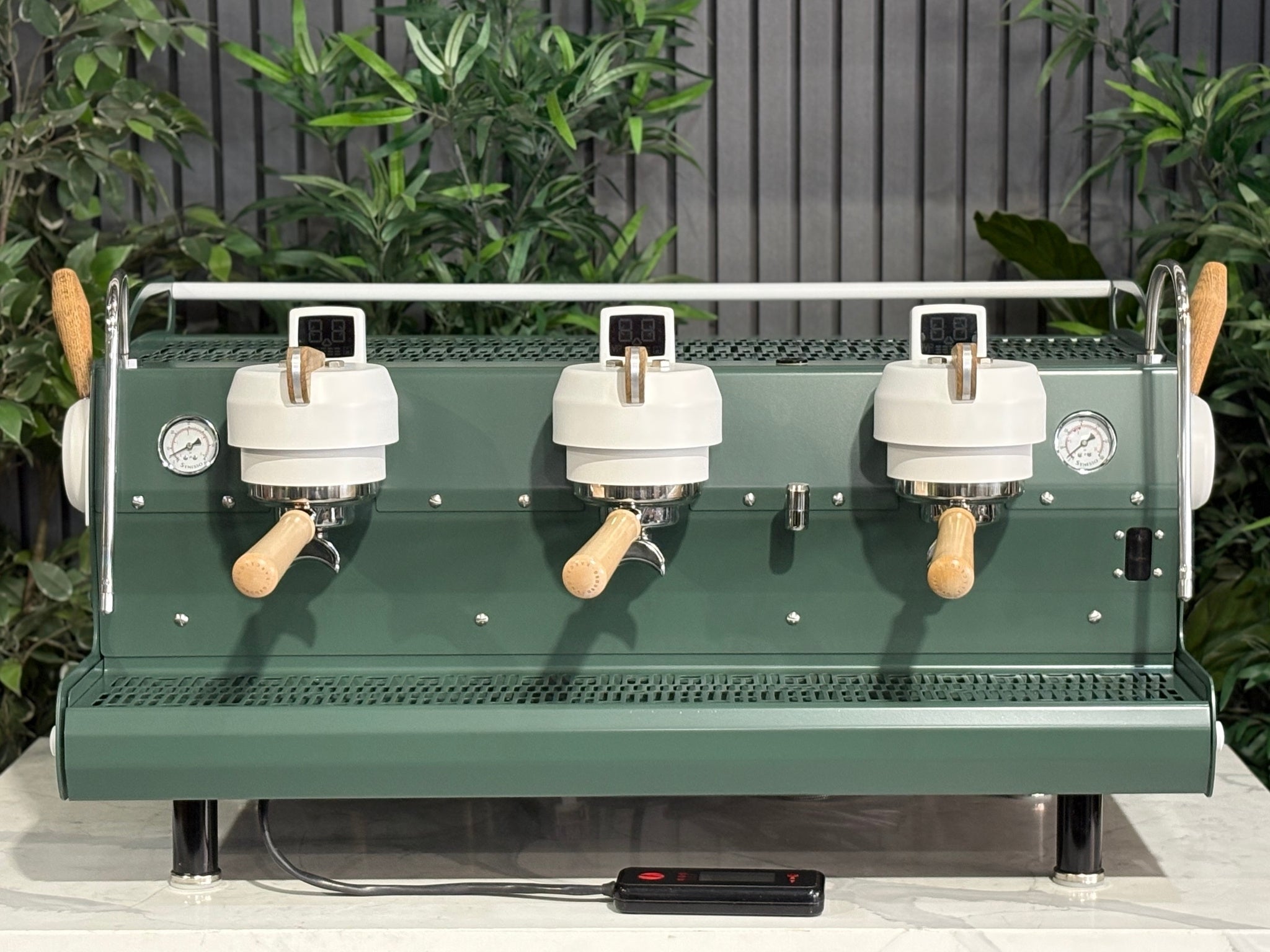 Synesso MVP 3 Group Coffee Machine Green, White & Timber - Used, front-on view highlighting the three group heads, dual cool touch steam wands with ergonomic levers, cup tray, programmable hot water tap, and digital shot timers, set on a marble bench with greenery behind.