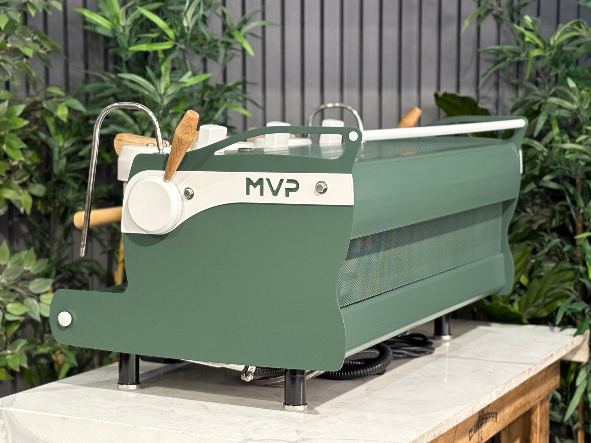 Synesso MVP 3 Group Coffee Machine Green, White & Timber - Used, side view showing green and white painted body, timber accents, dual steam wands with ergonomic lever handles, and cup tray, on a marble bench with greenery behind.