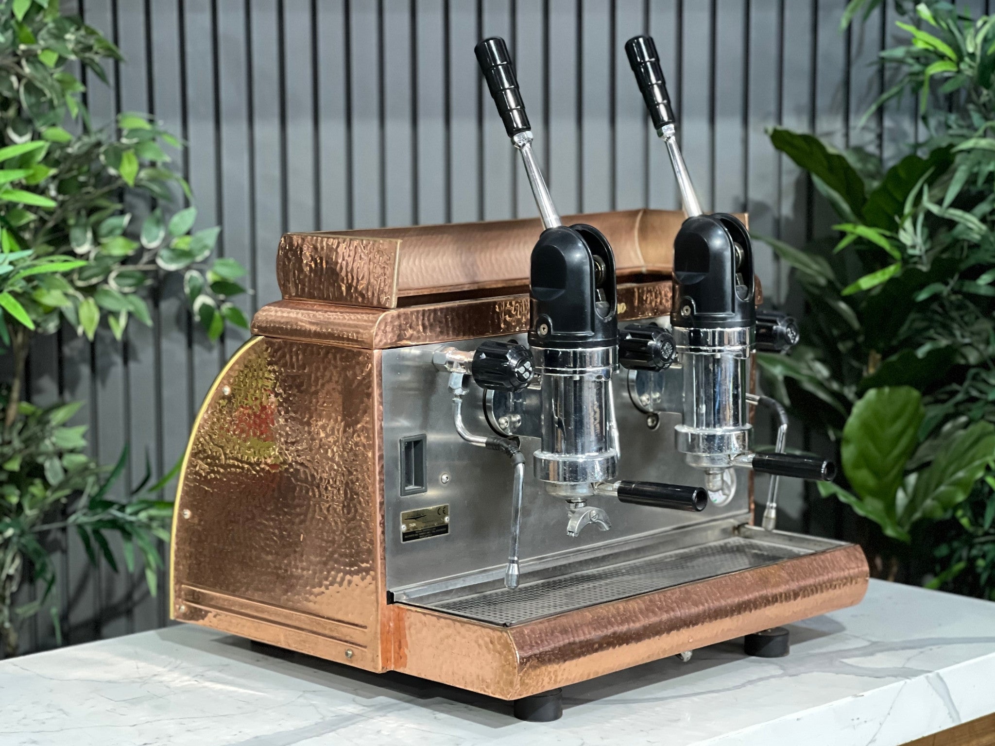 Victoria Arduino Athena Leva 2 Group Coffee Machine Bronze – Used espresso coffee machine shown at an angle on a marble bench with greenery in the background, highlighting its bronze body, dual group heads, lever arms, steam wands, and hot water tap.