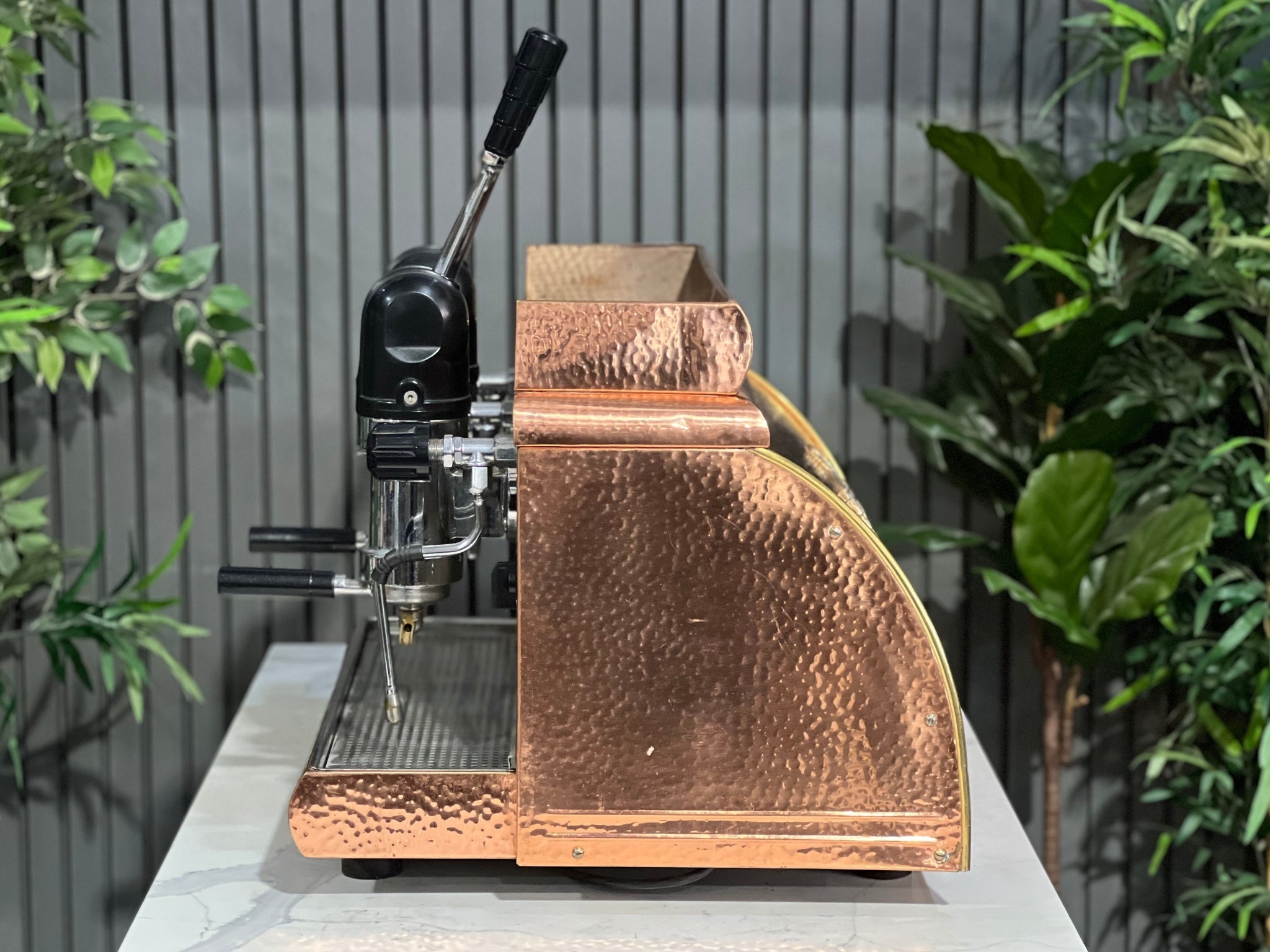 Victoria Arduino Athena Leva 2 Group Coffee Machine Bronze – Used espresso coffee machine side profile highlighting the lever mechanism, steam wand, and curvature of the bronze body against a marble bench backdrop.