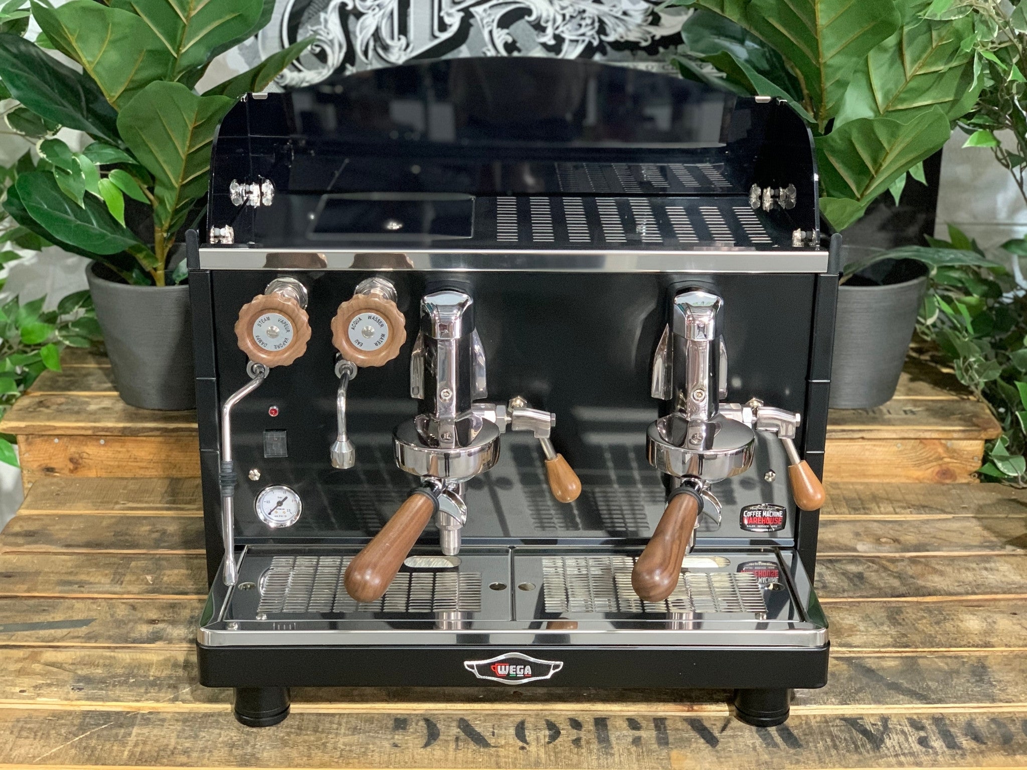 A brand new Wega Mini Nova Classic 2 Group Coffee Machine with a Matte Black finish & Timber Accents sitting on a wooden bench. There is greenery in the background. The image is taken from the front of the machine so that you can see the front of the machine, control panel, buttons and group heads.