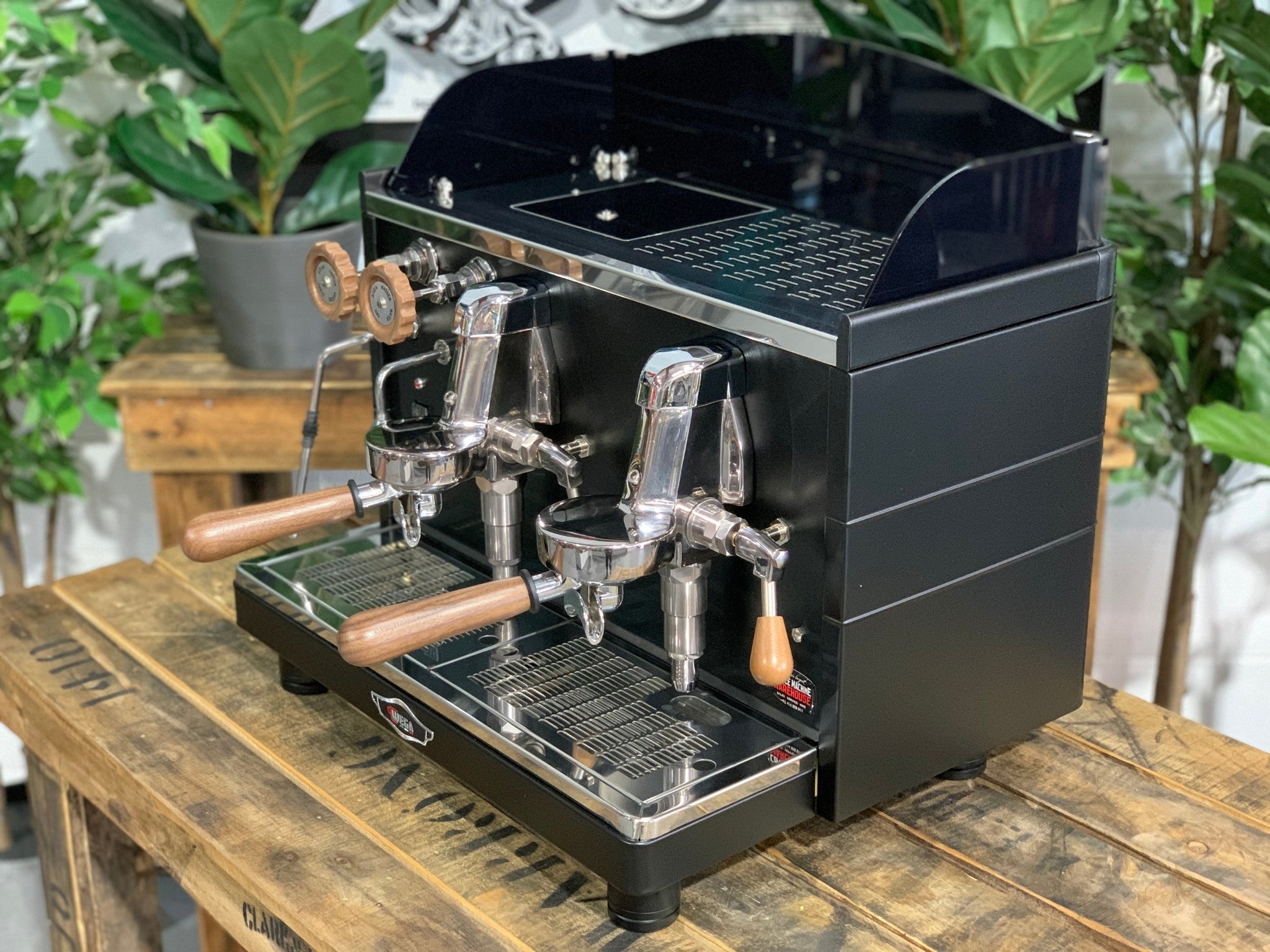 A brand new Wega Mini Nova Classic 2 Group Coffee Machine with a Matte Black finish & Timber Accents l is sitting on a wooden bench. There is greenery in the background. The image is taken from the front of the machine and the machine is sitting on an angle so that you can see the front of the machine and the side.