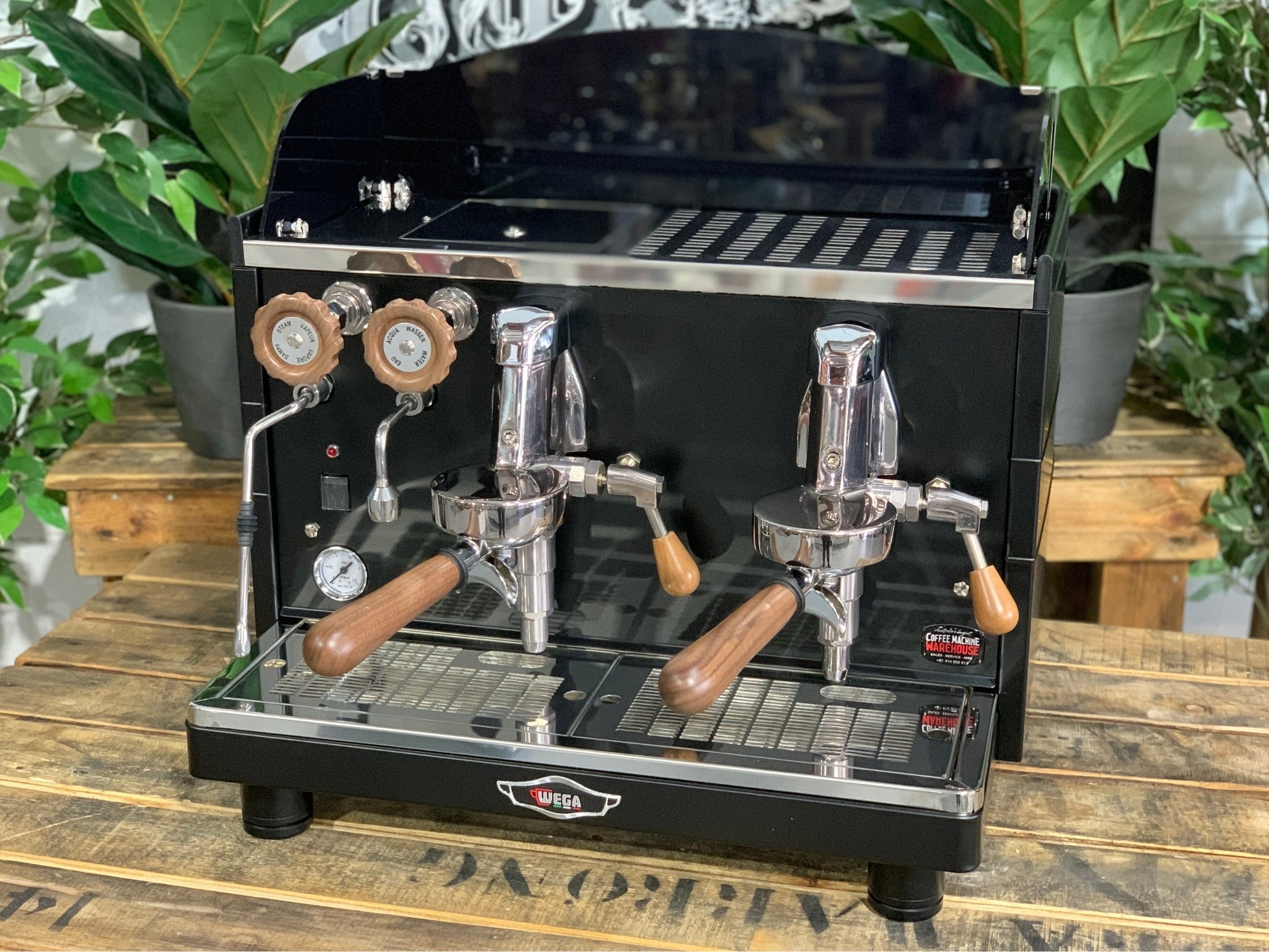 A brand new Wega Mini Nova Classic 2 Group Coffee Machine with a Matte Black finish & Timber Accents sitting on a wooden bench. There is greenery in the background. The image is taken from the front of the machine so that you can see the front of the machine, control panel, buttons and group heads.